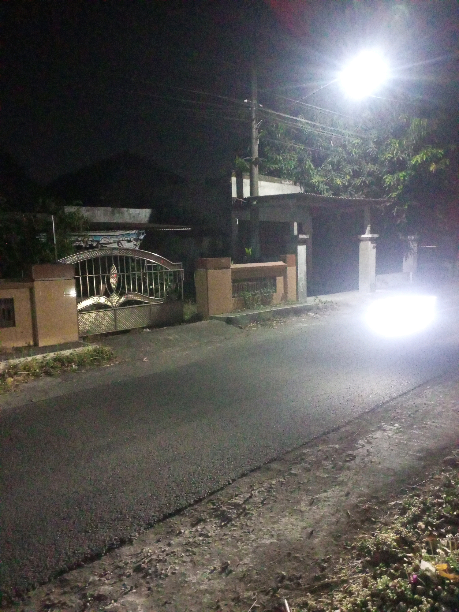 The image captures a nighttime street scene in the city of Ngasem, Indonesia. A bright street lamp illuminates the asphalt road, which appears to be somewhat uneven. On the left side of the road, there is a decorative metal gate with ornate patterns, flanked by two stone pillars. Beyond the gate, a house or building is partially visible, with a dark roof. Trees with dense foliage are present, particularly on the right side of the street, behind a building with a tiled roof and pillars. A bright glare from what appears to be vehicle headlights dominates the right side of the road, obscuring details of any approaching vehicle. The roadside on the right is overgrown with vegetation and scattered debris. The overall atmosphere is dark and somewhat hazy due to the artificial lighting and potential dust or mist in the air.
