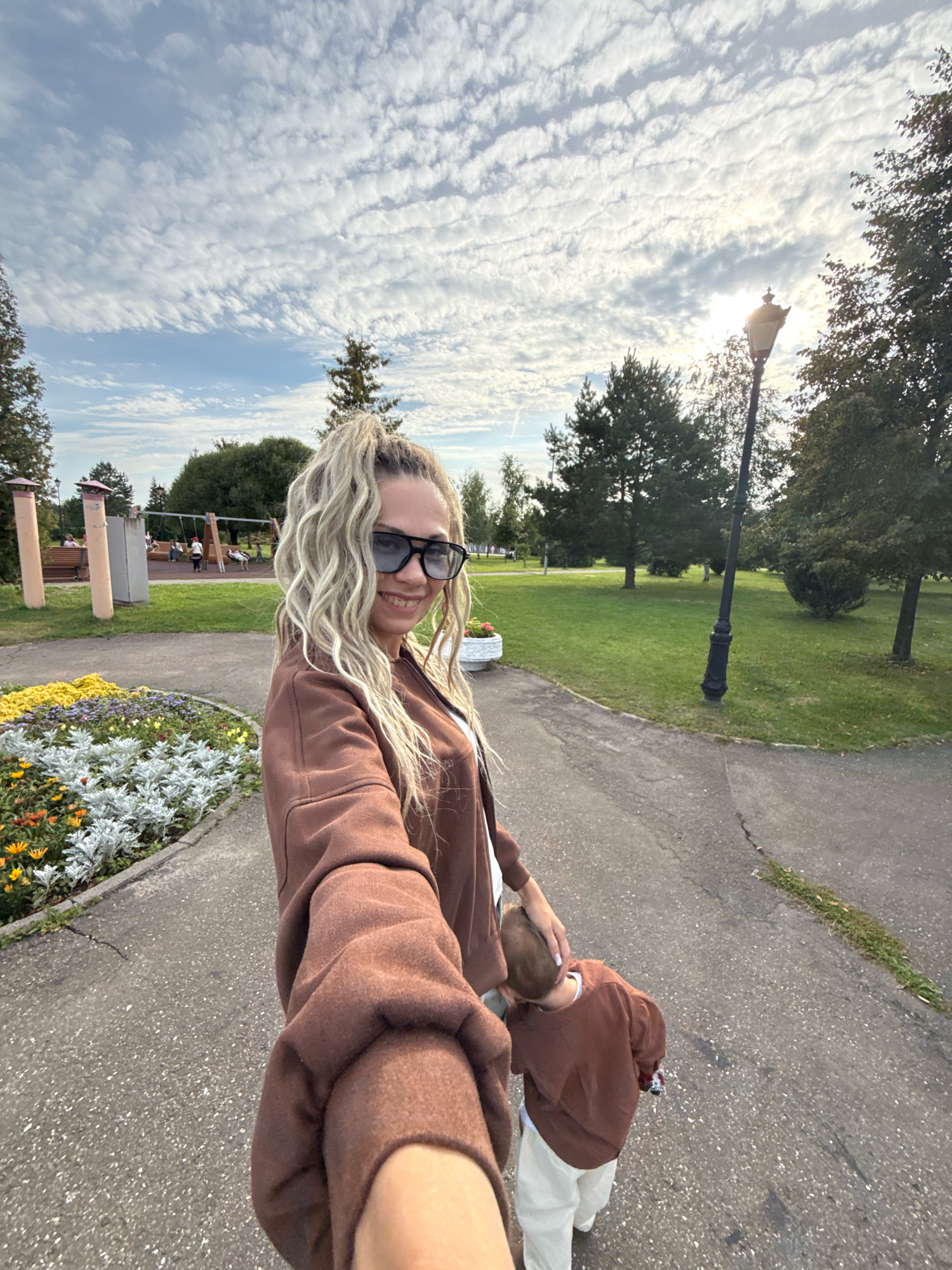 The image depicts a woman with blonde, curly hair pulled into a high ponytail, wearing sunglasses and a brown jacket. She is taking a selfie while walking on a paved path in a park. To her left, there is a flower bed with colorful blooms and some silver-leafed plants. In the background, a playground with swings and children can be seen, along with trees and a street lamp. A young child, wearing a brown sweatshirt and light-colored pants, is walking ahead of the woman, holding her hand. The scene appears to be in a city park, with a slightly overcast sky. The location is identified as Zhukovo, Russia.