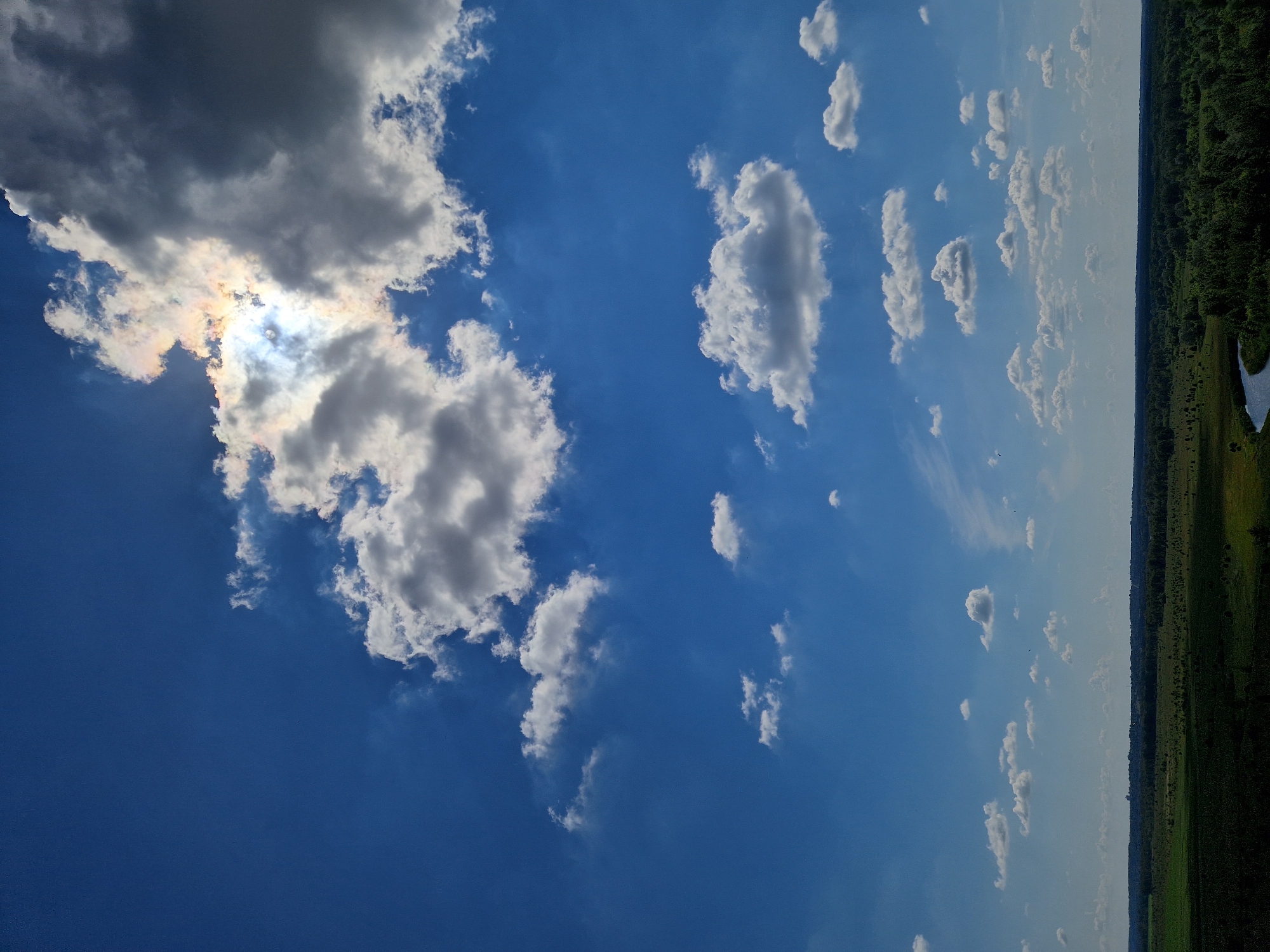 The image captures a panoramic view of the Ustyvytsia, Ukraine landscape on a sunny day. The sky is a vibrant blue, dotted with numerous white and gray cumulus clouds. The sun is partially obscured by a large cloud formation in the upper right quadrant, creating a radiant halo effect with hints of iridescence. Below the sky, a vast expanse of green fields stretches to the horizon, interspersed with dark green forests. A winding river with dark water snakes through the landscape, reflecting the blue of the sky. The overall scene depicts a tranquil natural environment.