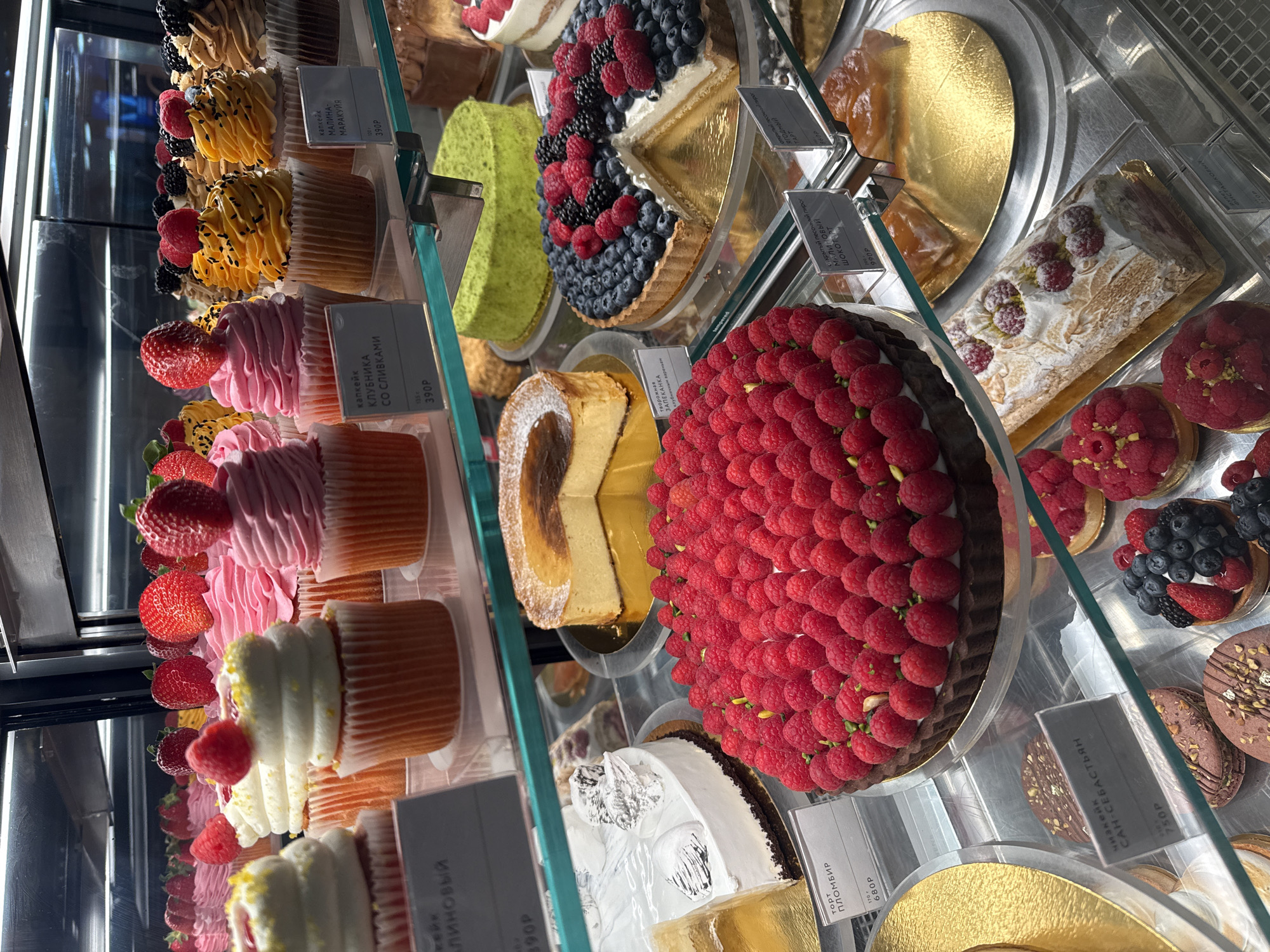 A display case in an establishment in Unknown, Unknown, presents a variety of desserts. Cupcakes topped with strawberries, pink frosting, or white frosting line the left side. Cakes, including a raspberry tart, a cheesecake, and a fruit tart with blueberries and raspberries, occupy the center and right sections. Small signs, in Russian, with prices are visible in front of some items.