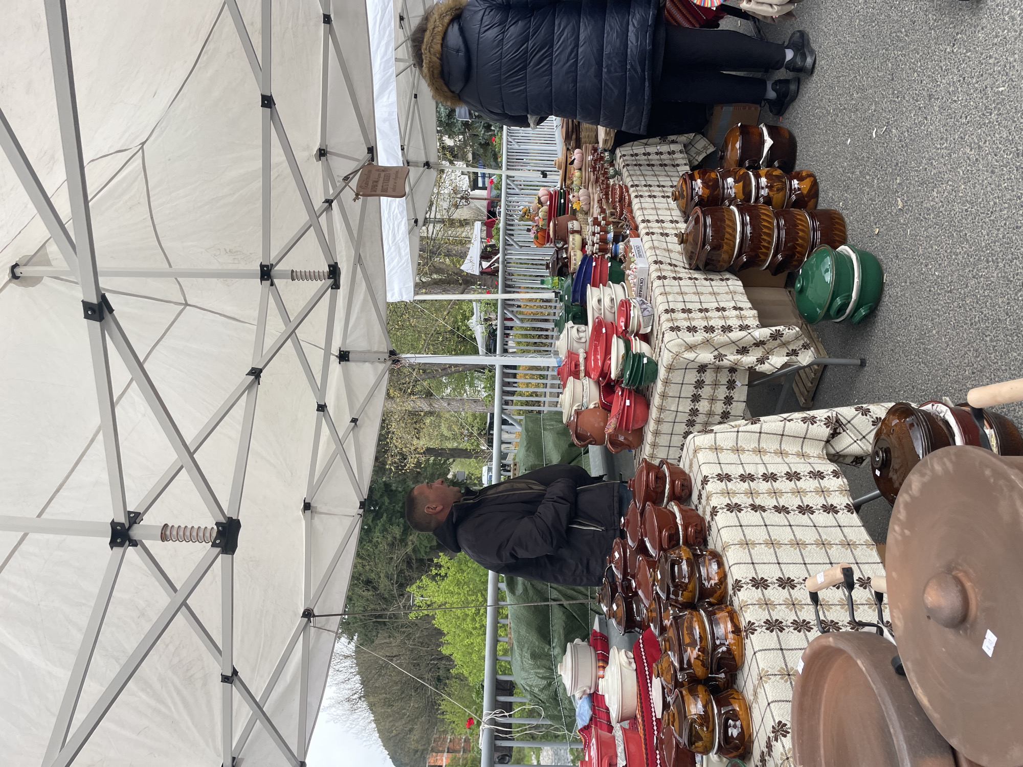 This outdoor scene captured in Zlatograd, Bulgaria, presents a market stall under a white canopy tent, displaying an array of pottery items. Predominantly, the pottery consists of brown glazed pots of various sizes. There are also some other colors like red, white and green. A man wearing a dark jacket is looking at the pottery, while another person in a dark puffer jacket stands at the corner of the table. The pottery sits on tables covered with beige and brown patterned tablecloths. In the background, greenery and buildings are visible. The scene suggests a local market or fair with crafts for sale.