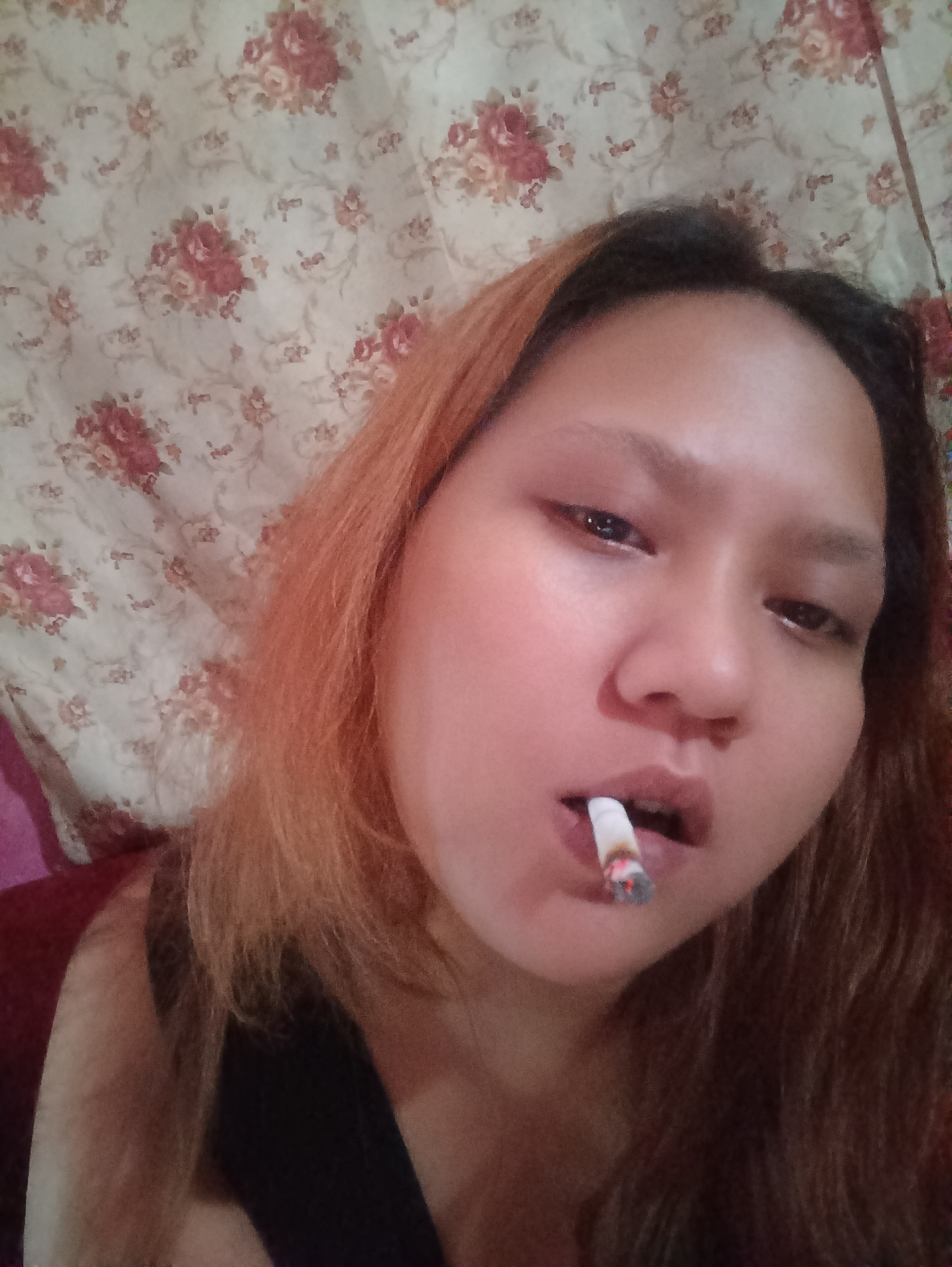 The image shows a close-up of a young woman with reddish-brown hair and light-colored skin, holding a lit cigarette in her mouth. She is looking directly at the camera with her mouth slightly open around the cigarette. The background is a floral patterned wallpaper. The lighting is soft and appears to be coming from the front. The woman is wearing a dark-colored top. There is no visible indication of the location in the city of Depok, Indonesia, or any specific environmental details that would help identify the scene as being from that city.