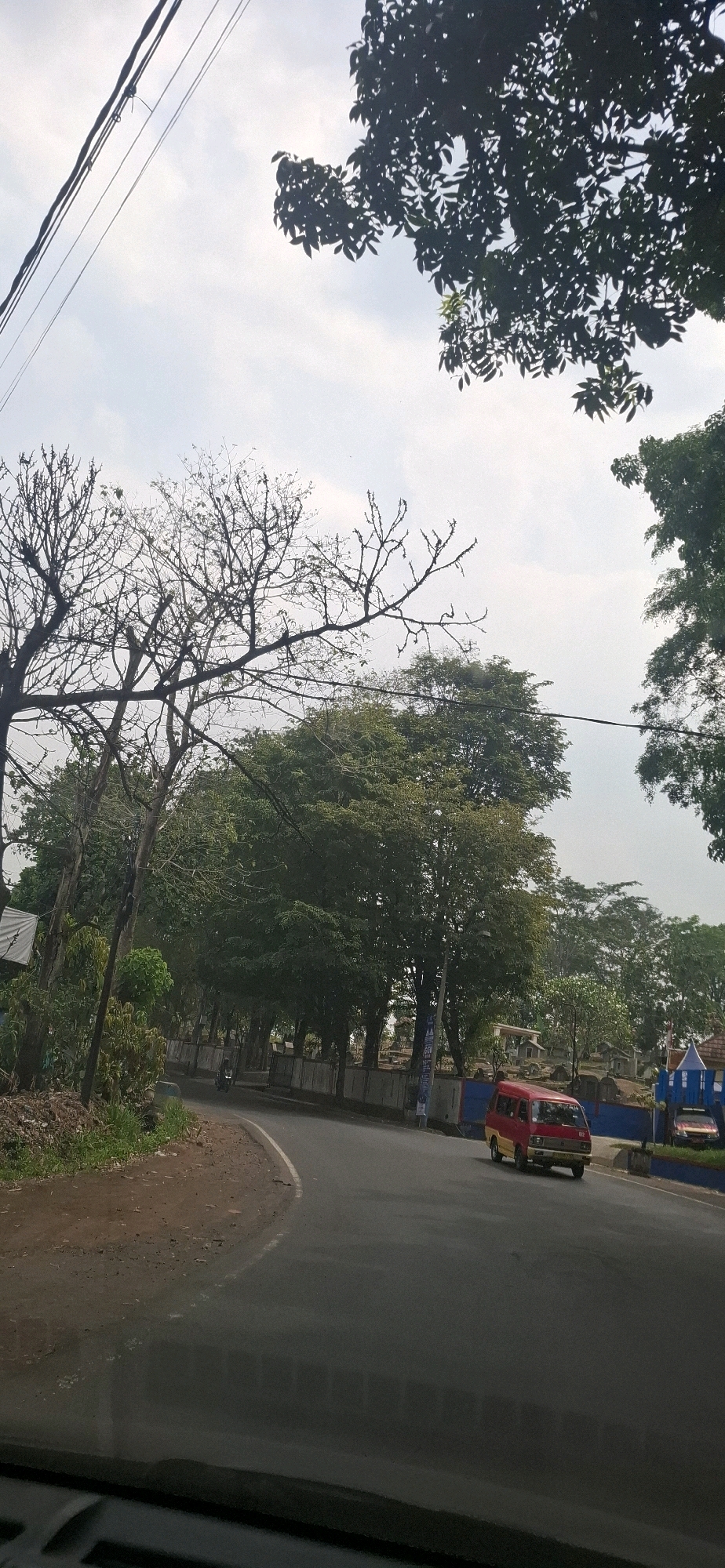 This is a street-level view looking forward from a vehicle, capturing a road curving to the left, lined with trees and some buildings. The sky is overcast, and overhead power lines are visible. On the right side of the road, a red and yellow van is driving away from the viewer. Further down the road, a few other vehicles and small buildings are present. The left side of the road features a dirt shoulder with grass and foliage, and the edge of the road is marked with a white line. The image appears to be taken in Sirnagalih, Indonesia, given the context.