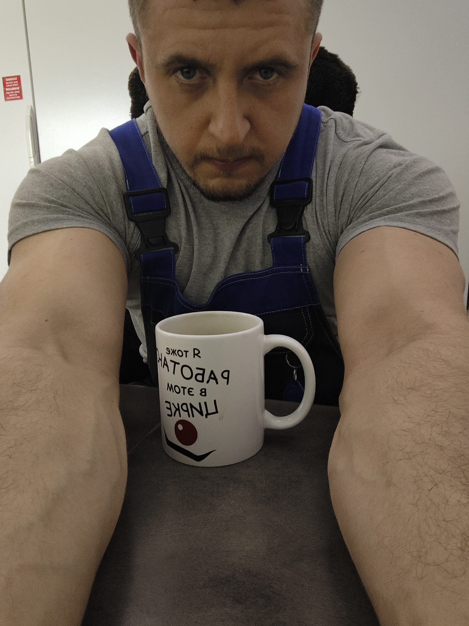 A low-angle, close-up shot features a man with short brown hair, blue eyes, and a light beard stubble. He is wearing a gray t-shirt and blue overalls with black straps. His muscular arms are visible in the foreground, leading toward a white mug with backward writing. The mug rests on a gray surface. The background includes a white wall.