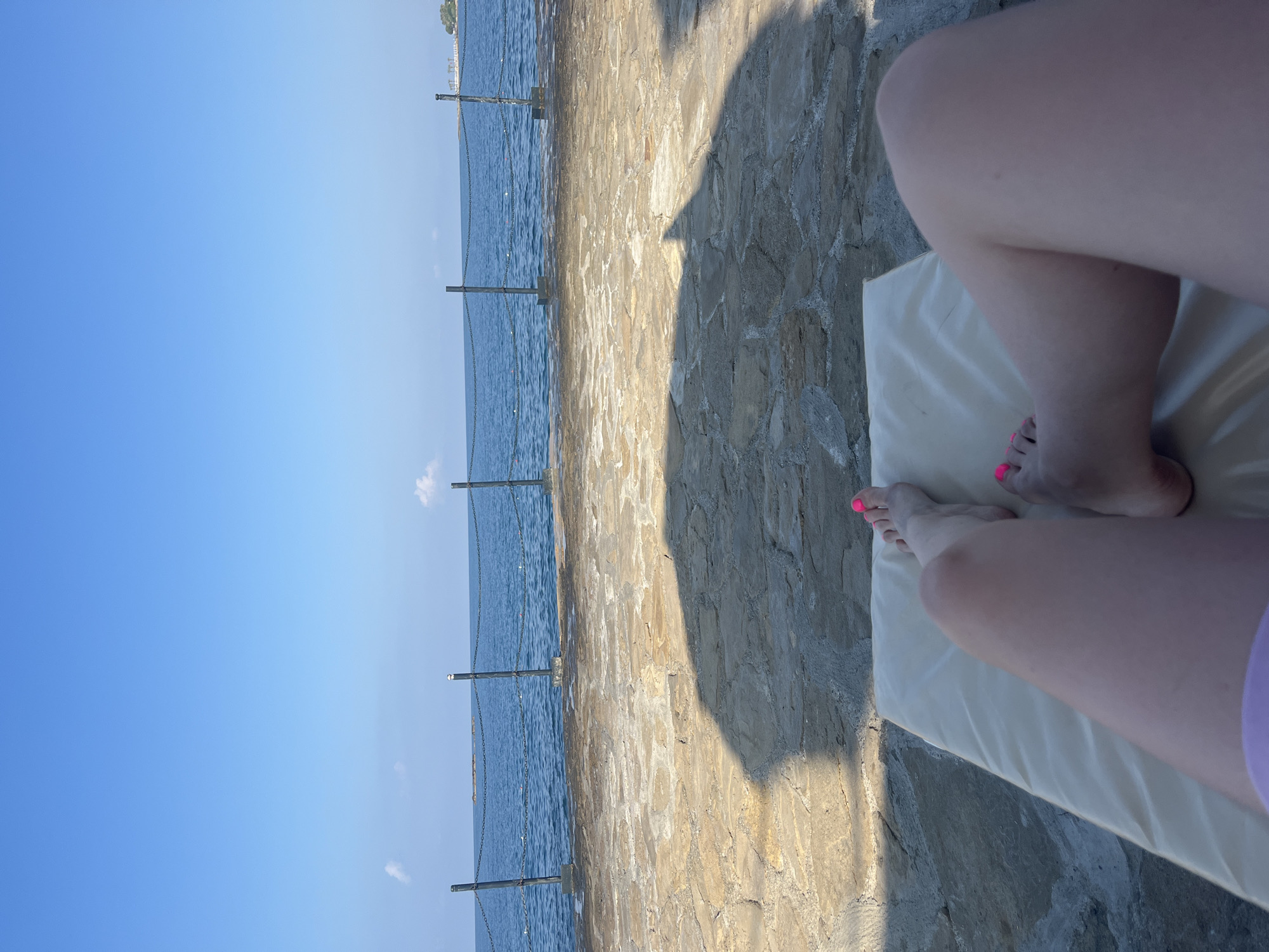 A first-person perspective shot shows a person relaxing on a recliner chair by the sea. The person's legs are visible, with bright pink nail polish on their toes. The recliner is placed on a stone patio, with a shadow cast by an unseen object. Beyond the patio, the blue sea stretches out to the horizon, with a few small islands in the distance. A chain-link fence runs along the edge of the patio, separating it from the water. The sky is a clear blue with a few white clouds. This scene is likely located in Alanya, Turkey, given the characteristic stone patios and the proximity to the Mediterranean Sea.