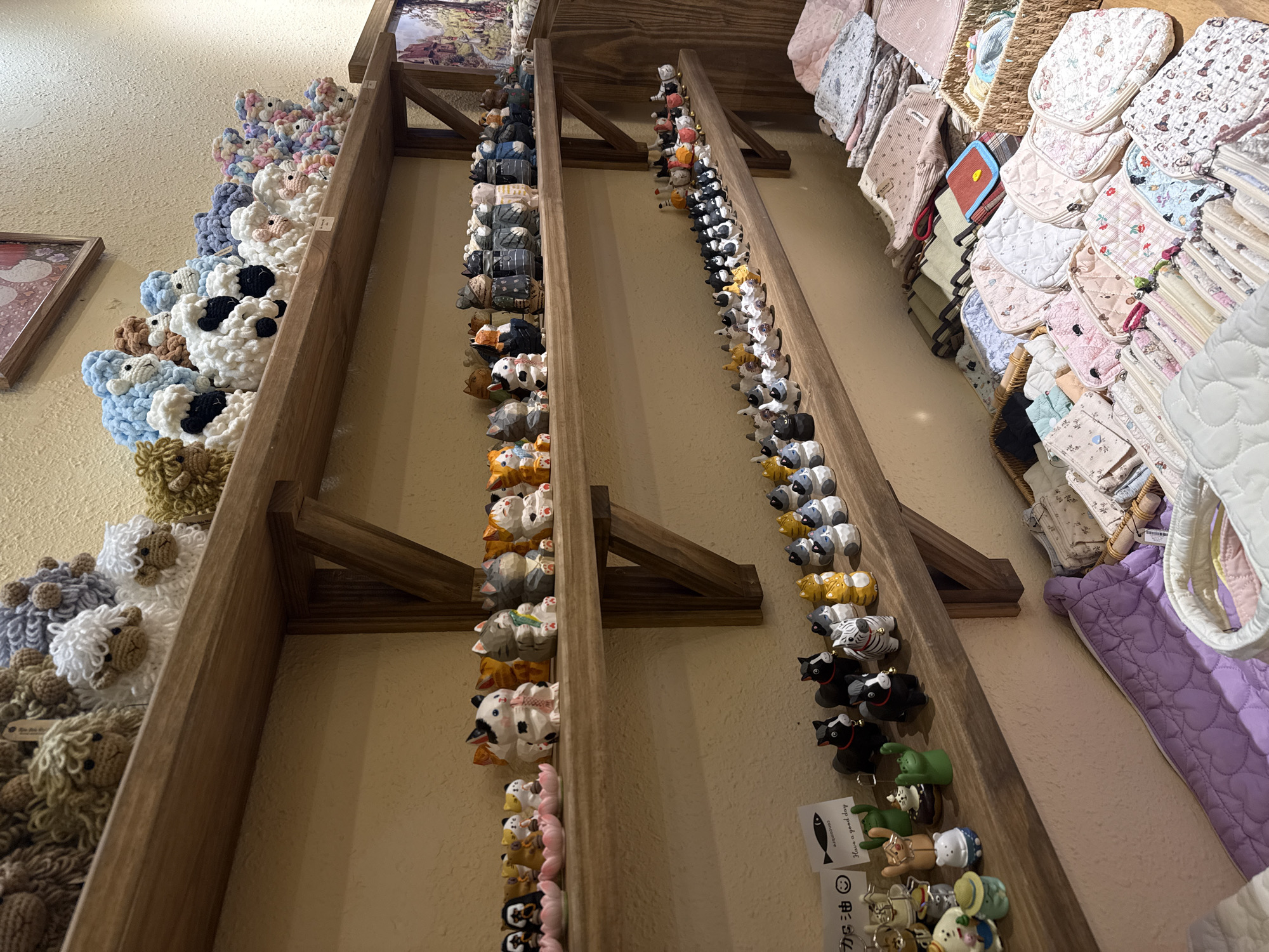 This image captures the interior of what appears to be a charming gift shop or artisan craft store in Da Lat, Vietnam, showcasing a variety of handcrafted items displayed on wooden shelves against a light-colored, textured wall. No people are visible in the scene.
On the left side, a collection of crocheted sheep dolls or plush toys are neatly arranged on a broad wooden shelf. These adorable sheep come in various yarn textures and colors, including white, black, light blue, pink, brown, and green, each with small faces and distinct curly "wool." A framed picture with a floral design hangs on the wall just below this shelf.
In the center of the image, two parallel, narrow wooden ledges hold an extensive array of small, painted wooden animal figurines. These intricate figures, appearing to be predominantly cats (including several maneki-neko style), raccoons, and other creatures, are lined up in long rows, showcasing a diverse range of colors and designs.
To the right, a section is dedicated to stacks of neatly folded fabric items, possibly small quilts, blankets, or decorative mats. These items display a variety of patterns, including florals, checkered designs, and cartoon motifs, in soft pastel colors. Some are housed in wicker baskets, while others are simply stacked on wooden shelves.
The lighting is soft and even, suggesting an indoor setting, likely during the daytime. Below the wooden figurines on the right, small white tags are visible. One clearly displays the English text "have a good day" next to a simple fish drawing. Below it, Japanese characters, "から油", are partially visible on another tag or keychain. The overall atmosphere conveyed is one of warmth, creativity, and artisanal craftsmanship.