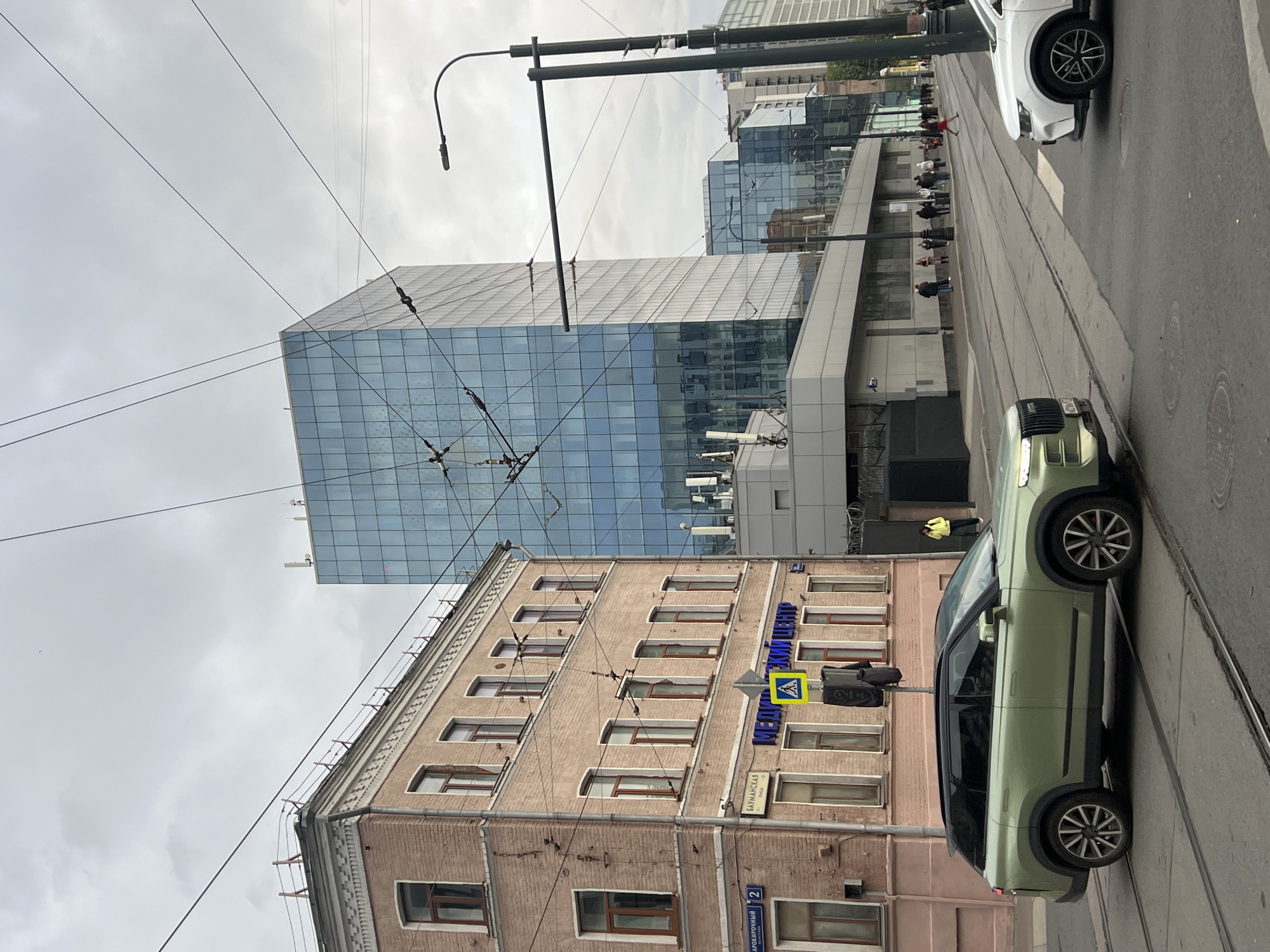 The image depicts a street scene in an urban environment. A modern glass-facade building rises in the background, adjacent to an older brick building with numerous windows. Power lines crisscross the sky above the buildings. Street signs are visible on the brick building, though the writing is not easily legible. A pedestrian crossing sign is also present. Two cars are positioned on the right side of the frame, on what appears to be a street with tram lines. People are present in the background near the glass building, seemingly queuing or waiting. The sky is overcast, suggesting a cloudy day. The overall impression is of a city street combining older and modern architecture, with typical urban elements such as traffic and pedestrian activity.