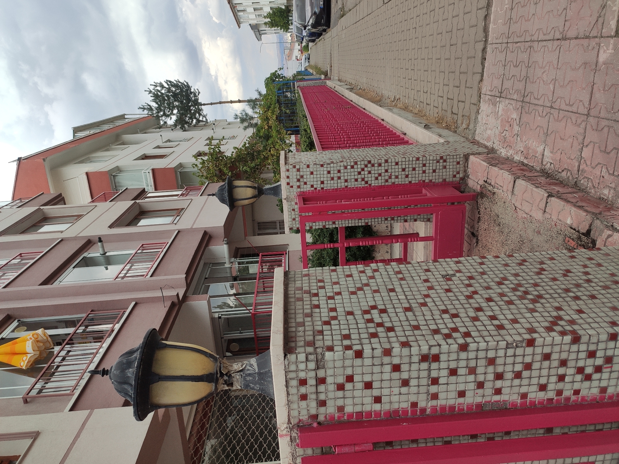 A street scene in Mamak, Turkey, captured from a low angle looking up. In the foreground, a tiled pillar with a black and yellow lantern is partially visible. A bright pink metal fence borders a walkway paved with interlocking bricks. Behind the fence, a building with a pink and white facade rises, featuring balconies with red railings and windows. Trees and more buildings are visible further down the street. Several cars are parked along the side of the road, and the sky is overcast.