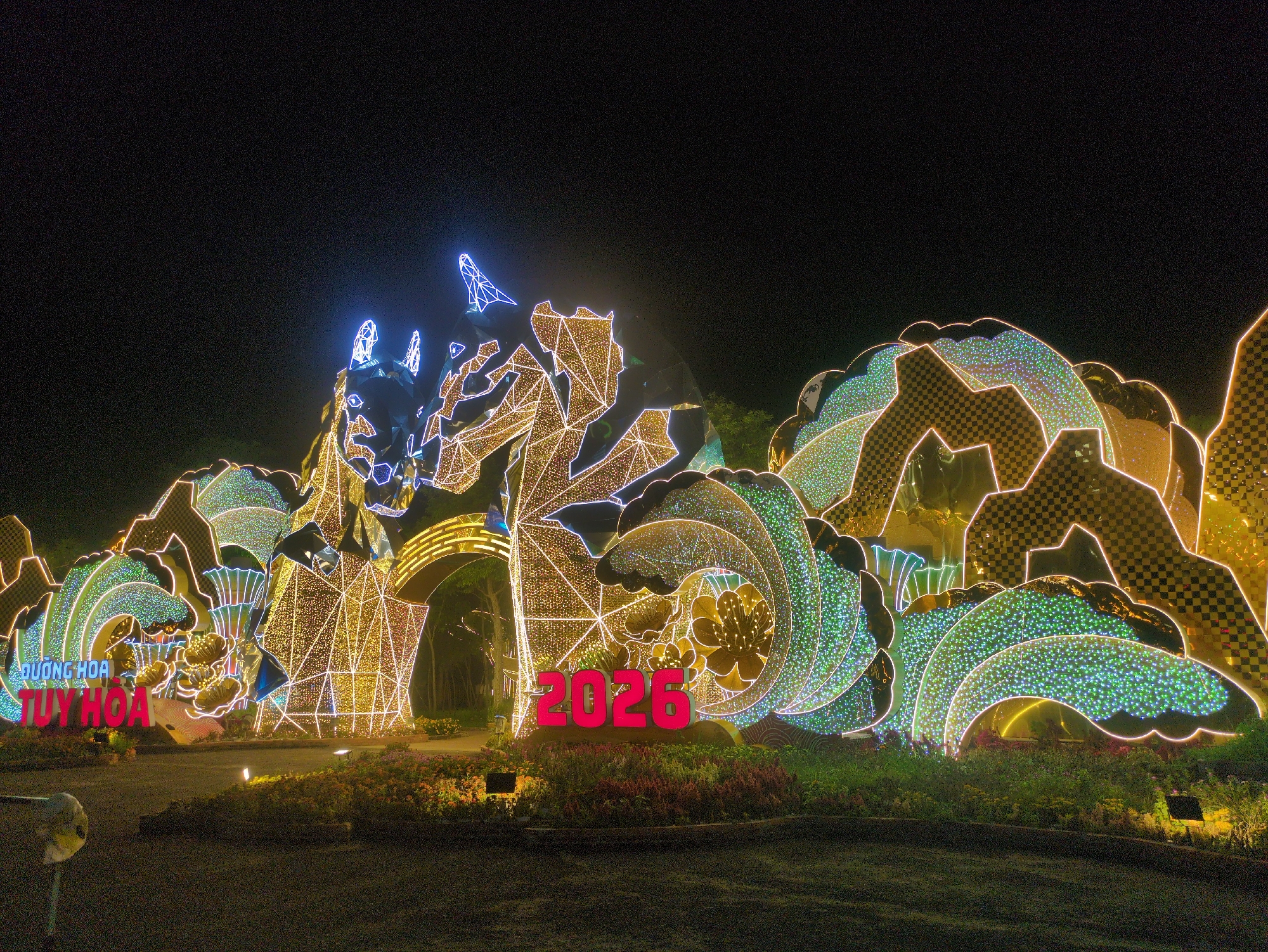 The image shows an illuminated display at night, likely in Tuy Hòa, Vietnam. The display features the year "2026" in large, red numerals. Behind it, a geometric structure resembling a dragon is illuminated with white and gold lights. To the sides, wave-like shapes are decorated with green and gold patterns. The display is situated in a garden or park, with flowers and greenery at the base. The sky above is dark. The phrase "Đường Hoa Tuy Hòa" is visible on the left side of the image.
