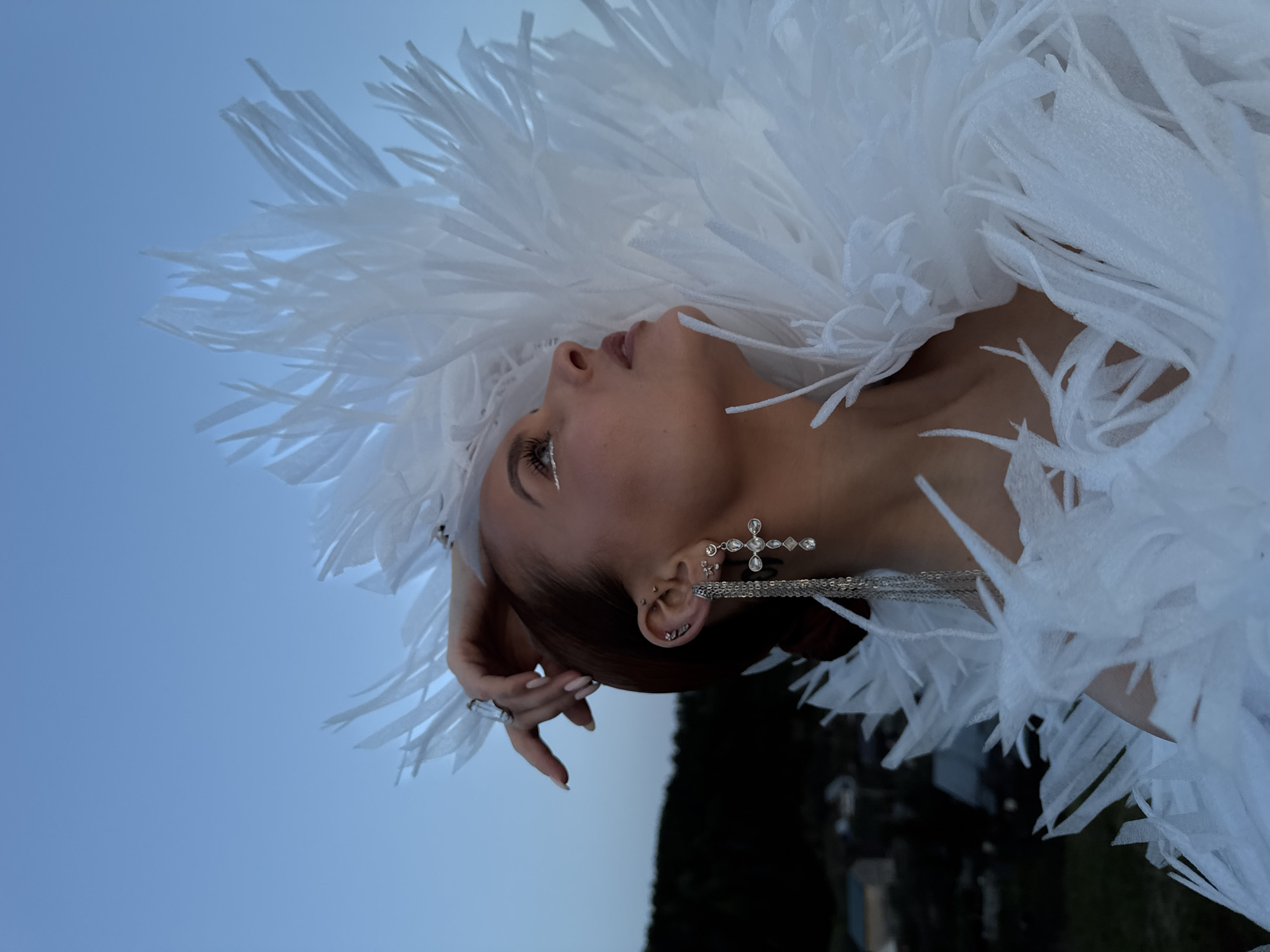 A woman with tan skin is photographed from a low angle, her face angled upward. She is adorned with a white, feathery garment around her head and shoulders. Her hair is pulled back, revealing multiple piercings in her ears, one of which is a large, cross-shaped earring. The woman is wearing a ring on one of her fingers, and she has a line of glitter-like makeup near her eye. The background consists of a blue sky and a blurry area, possibly trees, implying an outdoor location. The photo doesn't provide sufficient details to determine that it was taken in the city of Ольховка, Russia.