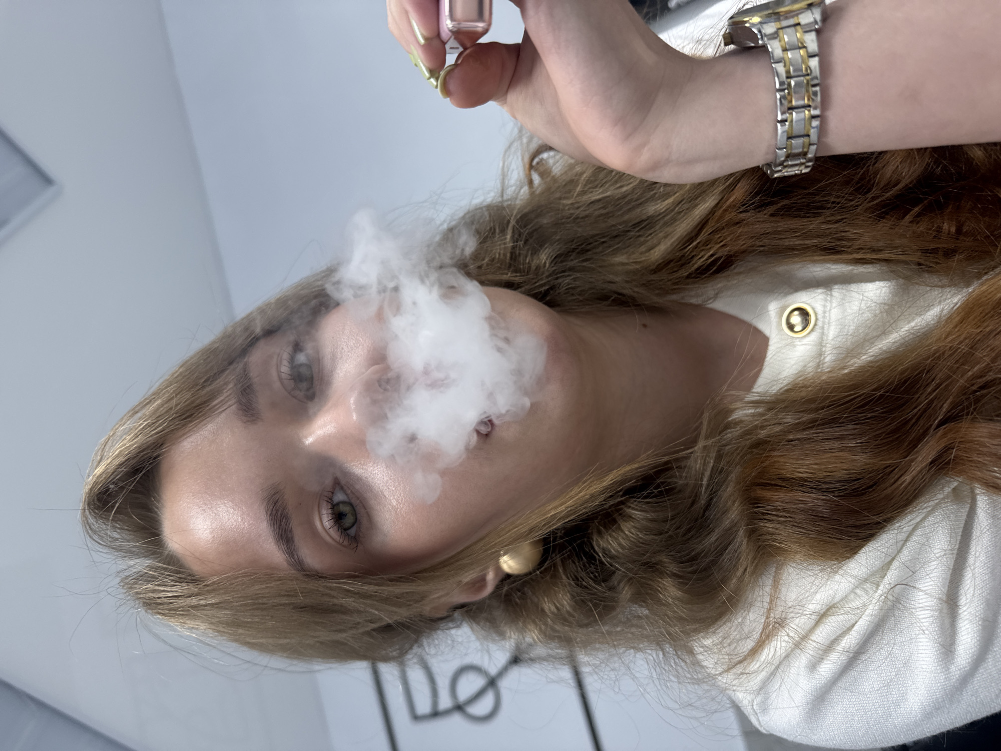 An image, rotated 90 degrees clockwise, captures a young woman looking directly at the viewer while exhaling a dense cloud of white smoke or vapor, which partially obscures her lower face. Her long, wavy, reddish-brown hair cascades around her head.
She is wearing a white garment, possibly a shirt or jacket, featuring a prominent gold button near her shoulder. On her right wrist, she sports a two-toned silver and gold metallic wristwatch. Her right hand, adorned with light green nail polish, holds a small, clear bottle with a pink cap, likely a vaping device or e-cigarette, which is the source of the visible vapor. A gold stud earring is visible in her right ear.
The background is a plain, light-colored (white or light grey) indoor wall or ceiling. A rectangular ventilation grate can be seen in the upper left corner of the background, suggesting an indoor, potentially commercial or office environment. The lighting is bright and uniform, indicating an indoor setting with artificial light. The woman's expression is neutral and direct. There is no visible text. The scene is located in Chaikovsky, Russia.