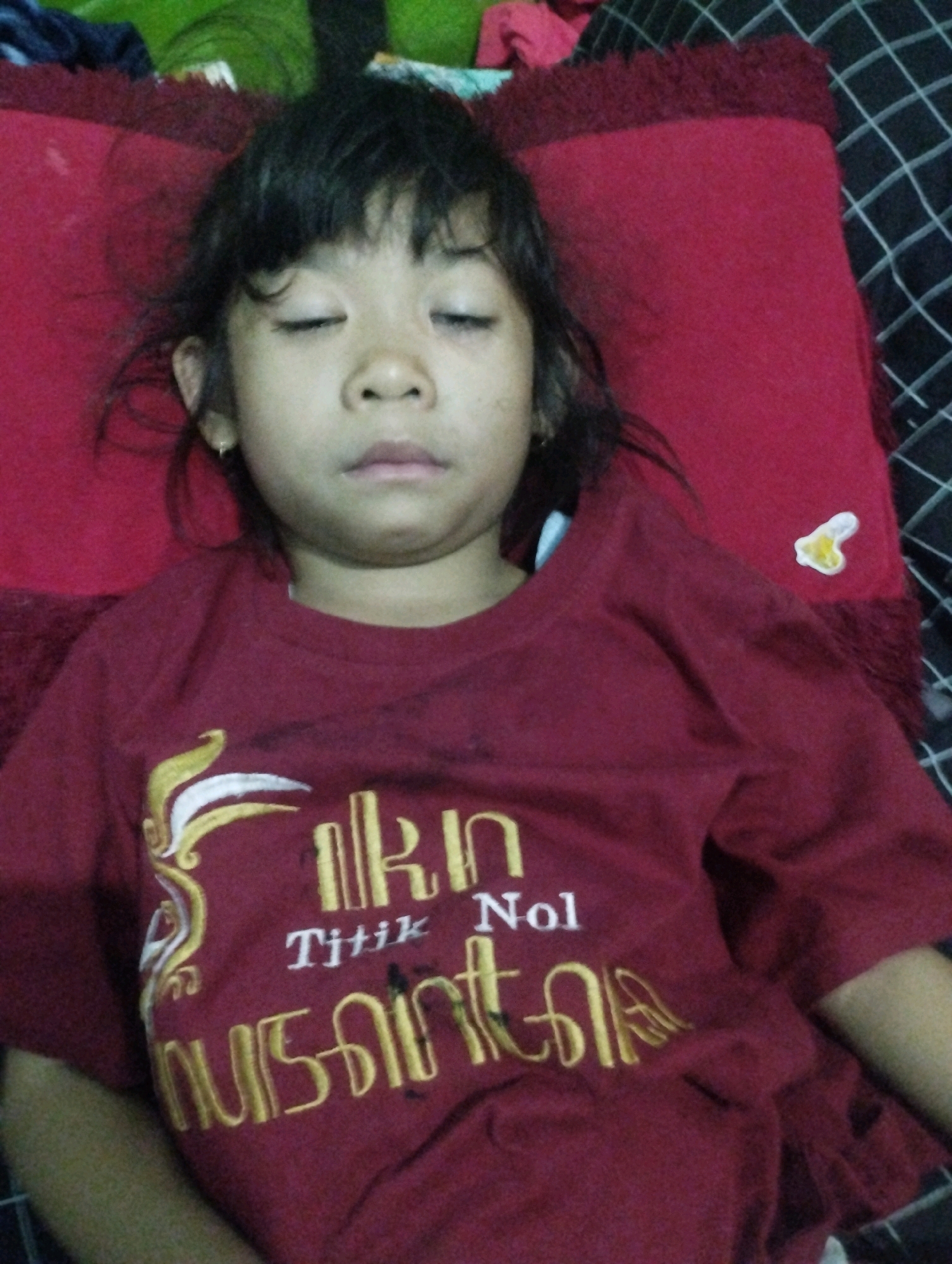 A young Asian girl with closed eyes and dark hair is lying down. She is wearing a maroon t-shirt with yellow and white embroidery on the front. The t-shirt has an emblem on the left chest and text on the right. The girl is on a red pillow which is on top of a black and white checkered surface. The image appears to be taken in Kemantren, Indonesia, likely in a bedroom or domestic setting.