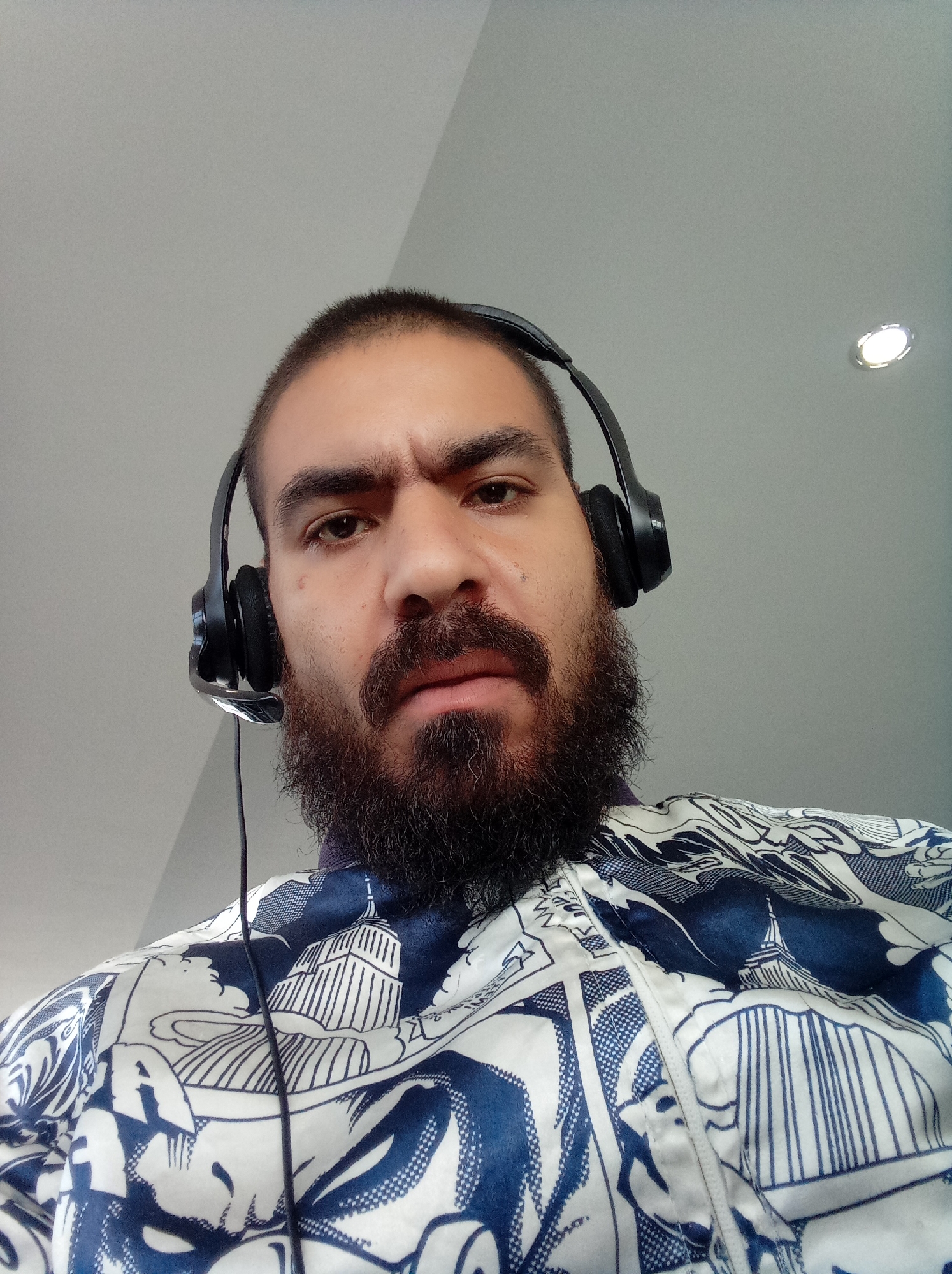 A medium, eye-level close-up shows a man with a dark beard and short dark hair wearing black headphones with a microphone attached. His expression appears stern, with furrowed brows. He is wearing a white and blue shirt with an artistic pattern depicting architecture. The background is a plain, light gray wall. A light fixture is visible in the upper right corner. The location cannot be determined from the image.