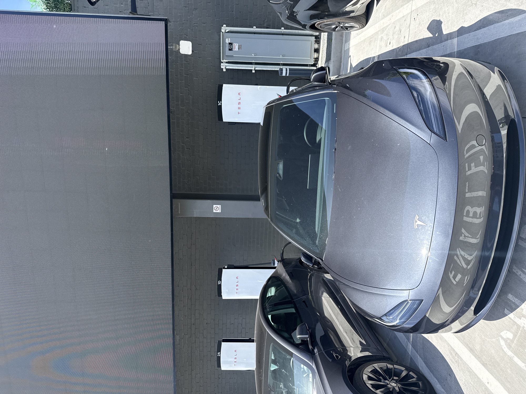 The image captures an outdoor scene at what appears to be a Tesla Supercharger station. Two gray Tesla cars are parked and charging, with one prominently featured in the foreground, partially obscuring the second. The background reveals multiple Tesla Supercharger units mounted on a dark gray brick wall, identified by their distinct white enclosures with the "TESLA" logo. Positioned above the charging units is electrical equipment in metal housings.
The setting is illuminated by daylight, casting shadows that define the parking space and surfaces of the cars. The environment is characterized by a paved parking area, and a portion of what may be a large outdoor display screen is visible on the left.
Based on the supercharger design and surrounding architectural details, this scene likely occurs within the city of West Hollywood, United States of America.