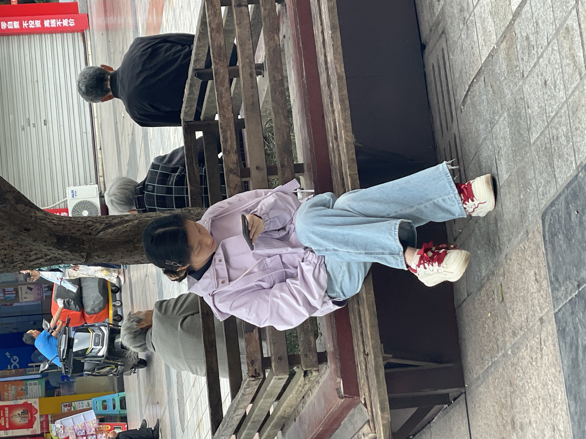 The image shows a street scene in Jiangjin, China. A young woman sits on a slatted wooden bench, wearing a light purple jacket, jeans, and white sneakers with red accents. She is looking down and holding a smartphone. Several elderly individuals are also sitting on the bench. One man is wearing a black jacket and another wears a grey shirt. Behind the bench, a man in a blue shirt drives a small electric vehicle. Further in the background are storefronts with red signage written in Chinese characters and miscellaneous street furniture. The ground is paved with grey tiles.