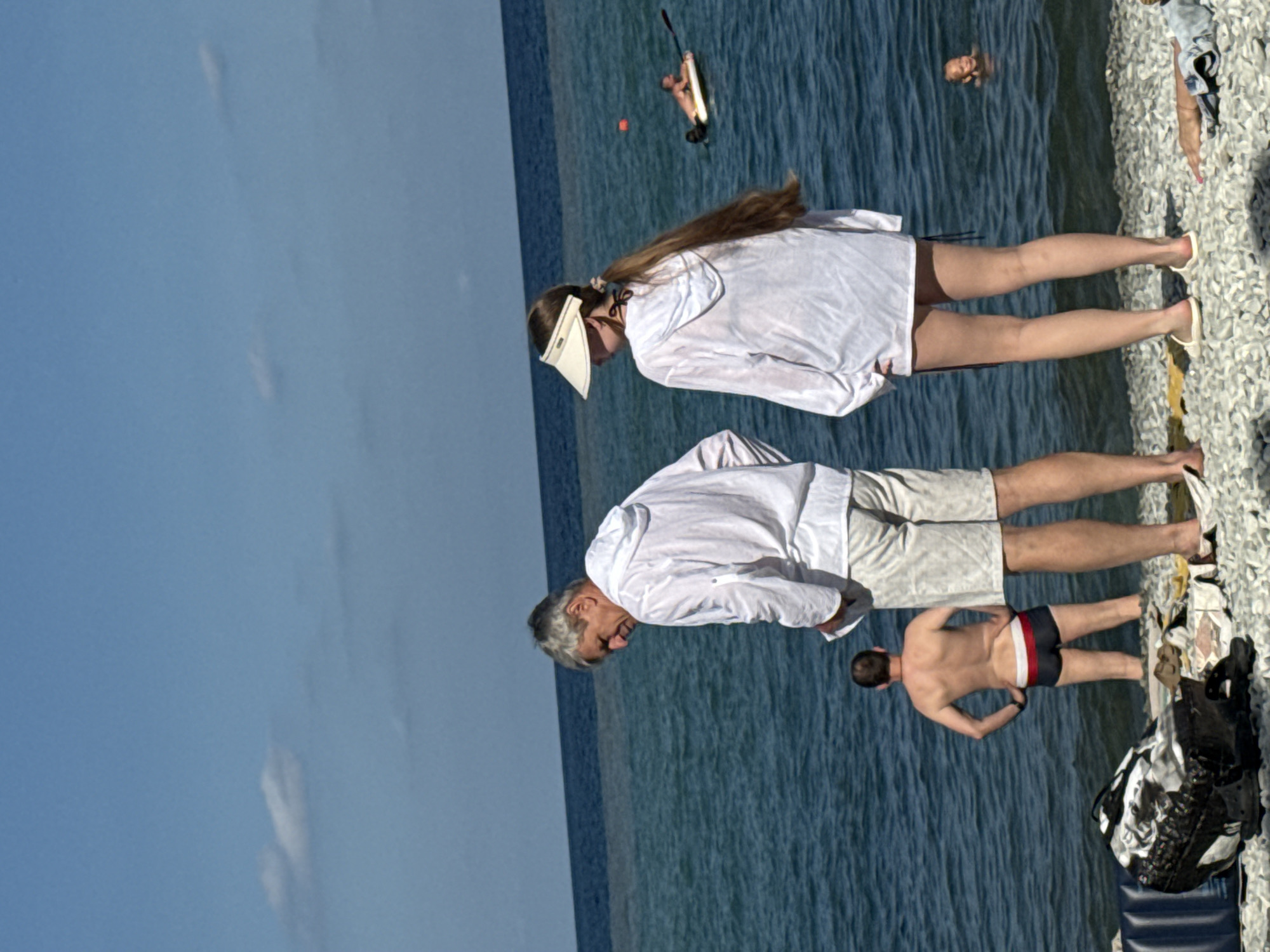 The image shows a seaside scene with several people. In the foreground, two individuals wearing white hooded shirts and shorts stand on a pebble beach, facing the water. To their left, a man in swim trunks stands in the shallow water. Further out in the sea, another man is seen paddling on a board. A child is also visible in the water in the background. The sky is blue with scattered clouds, and the sea is a deep blue. The setting appears to be a beach, possibly in the city of Partenit, Ukraine, given its coastal location.