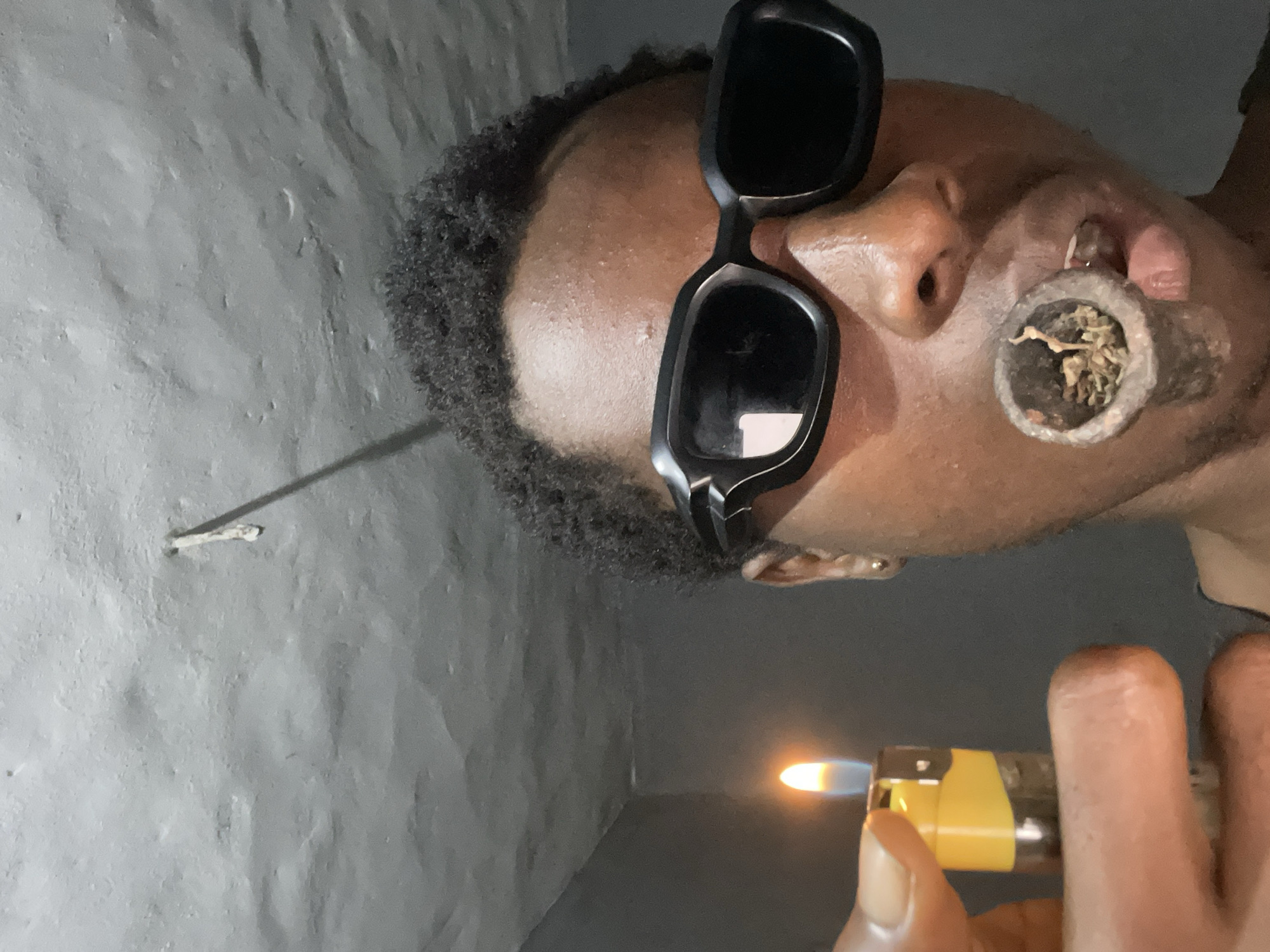 The image captures a close-up, portrait-style view of a person engaged in lighting a pipe. The subject is an individual with dark skin, seen in profile, wearing dark, rectangular sunglasses that obscure their eyes. Their textured, dark hair is visible on the side of their head.
A pipe, appearing to be made of wood or a natural material, is positioned near their mouth and contains dried plant matter. A hand, likely belonging to the person, holds a yellow and transparent disposable lighter with an active flame. The flame is directed towards the contents of the pipe, indicating the act of ignition for smoking.
The background is a textured, light gray wall, suggesting an indoor setting. A small, white object, possibly a nail or peg, protrudes from the wall in the upper left portion of the frame. The lighting in the scene appears to be artificial. No text is visible in the image. The location context provided is Onitsha, Nigeria.