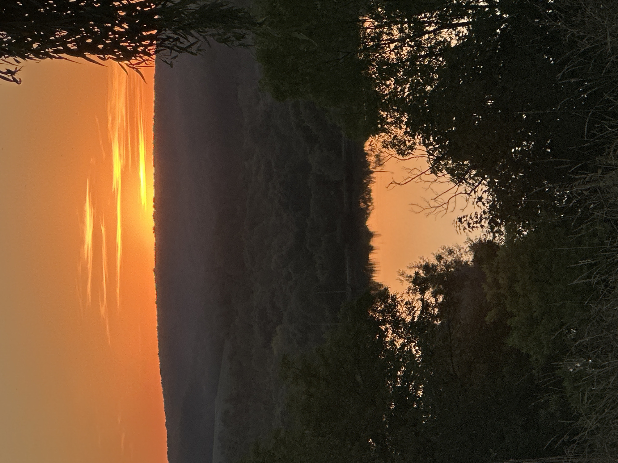 The image captures a tranquil sunset over a body of water, likely a lake or pond, located in or near Makiv, Ukraine. The sky is a vibrant gradient of orange and yellow, with thin, wispy clouds stretched horizontally across the horizon. Below the sky, a dark silhouette of rolling hills covered in dense forest forms the backdrop. In the foreground, the calm water reflects the colorful sky, creating a mirror image of the sunset. Trees frame the scene on both sides, their dark foliage contrasting with the bright sky and water. The overall impression is one of peacefulness and natural beauty, characteristic of a rural landscape at dusk.