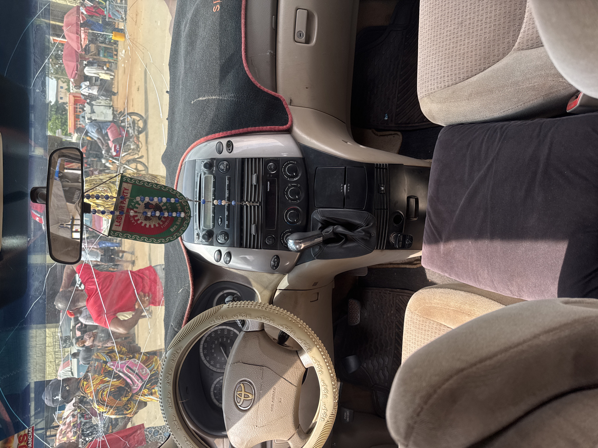 The image is taken from inside a vehicle, looking out through a cracked windshield at a bustling street scene in Orji, Nigeria. The dashboard and steering wheel are visible in the foreground, with a beige interior. A steering wheel cover with Arabic script is present. A car mat is visible on the floor.
Outside the vehicle, a group of people are gathered. One person in the foreground to the left wears a colorful patterned shirt and a pink fanny pack. Another person in the center is wearing a red t-shirt. There are market stalls with red umbrellas and various goods displayed. A motorcycle is visible in the middle ground. The environment appears to be a busy outdoor market or street. A "LABOUR PARTY" banner is hanging from the rearview mirror.