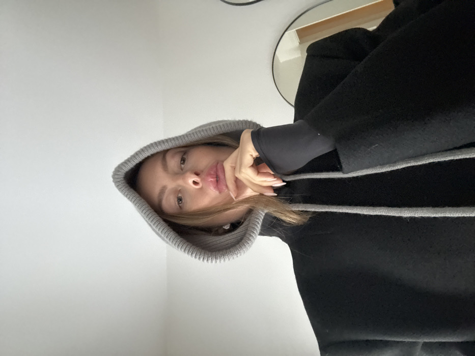 A low-angle, eye-level indoor shot depicts a female subject from the chest up, positioned centrally against a plain white wall. She wears a grey ribbed knit hood with two grey drawstrings, layered over a black high-collared garment. Her outer garment is a black coat or jacket, from which a black long-sleeved undergarment with a thumbhole extends on her right arm. Her light-skinned right hand, with light-colored fingernails, is positioned with fingers splayed against her chin, slightly obscuring her mouth which appears pursed. Her eyes are directed towards the camera. A small, reflective earring is visible on her left ear. To the right, a portion of an oval or circular mirror reflects a light-colored interior surface. The location is an unspecified indoor setting in Unknown, Unknown.