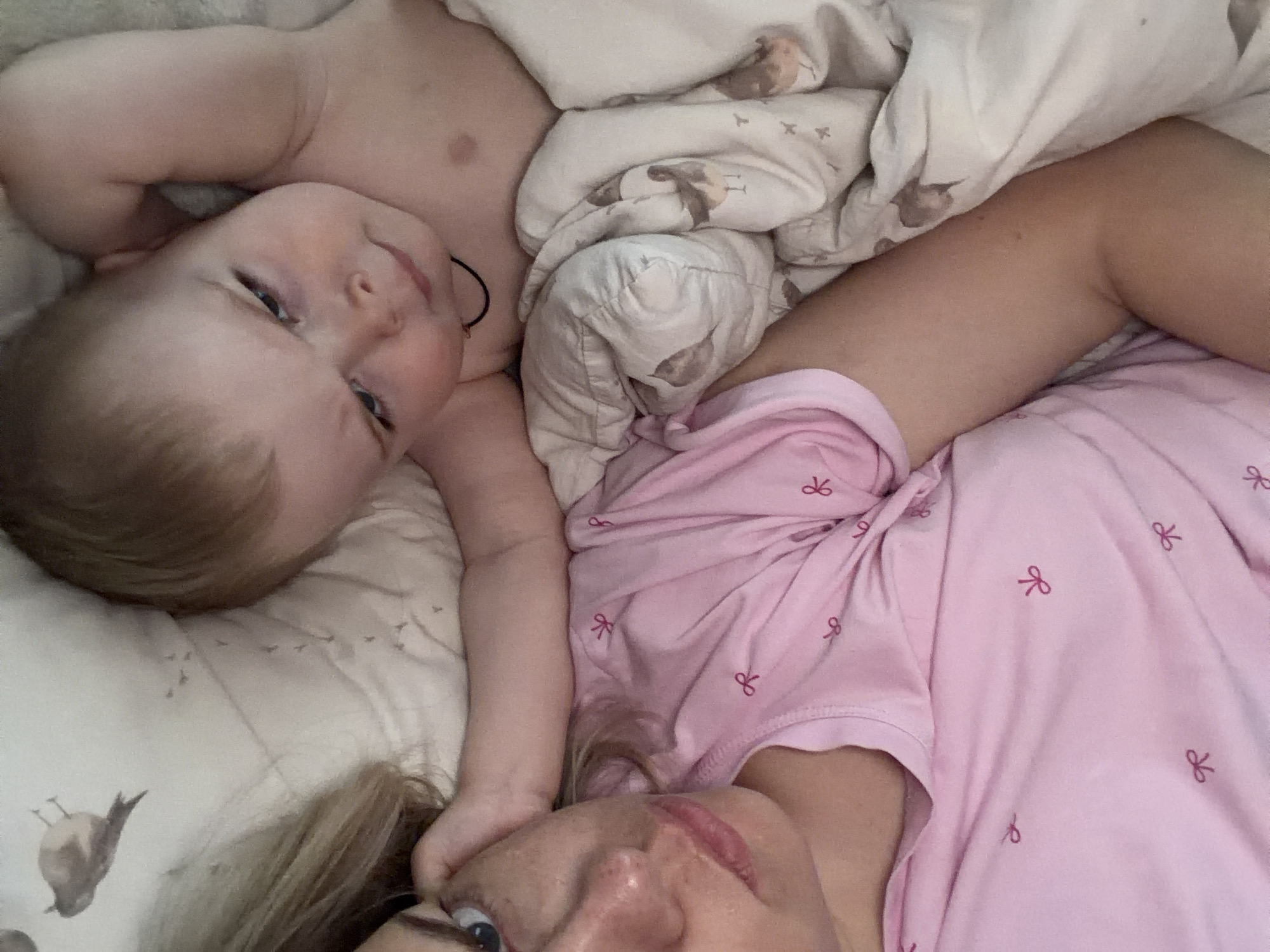 A photograph depicts a woman and a baby lying down, presumably in a bed. The baby is positioned near the top left of the frame, looking towards the viewer. The woman is in the lower right corner, with part of her face visible. She is wearing a pink shirt with a bow pattern. The bed is covered in a white blanket with a bird print. The image appears to be taken from an overhead perspective. There is no way to determine the location of the scene in Votkinsk, Russia, without additional information.