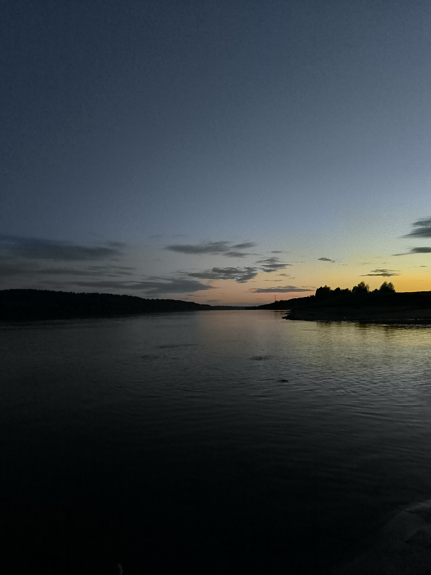 The image depicts a tranquil scene of a river at dusk. The sky transitions from a deep blue at the top to hues of orange and yellow near the horizon, indicating the setting sun. Clouds are scattered across the sky, with some appearing darker and more defined than others. On the left side of the image, a dark, silhouetted treeline forms a low ridge along the riverbank. The river itself is dark and reflects the dim light of the sky, with subtle ripples on its surface suggesting a gentle current. On the right side, another silhouetted treeline is visible, with the land sloping down towards the water. The overall atmosphere is serene and peaceful, characteristic of a quiet evening by the water. The location is specified as Zagotzerno, Russia.