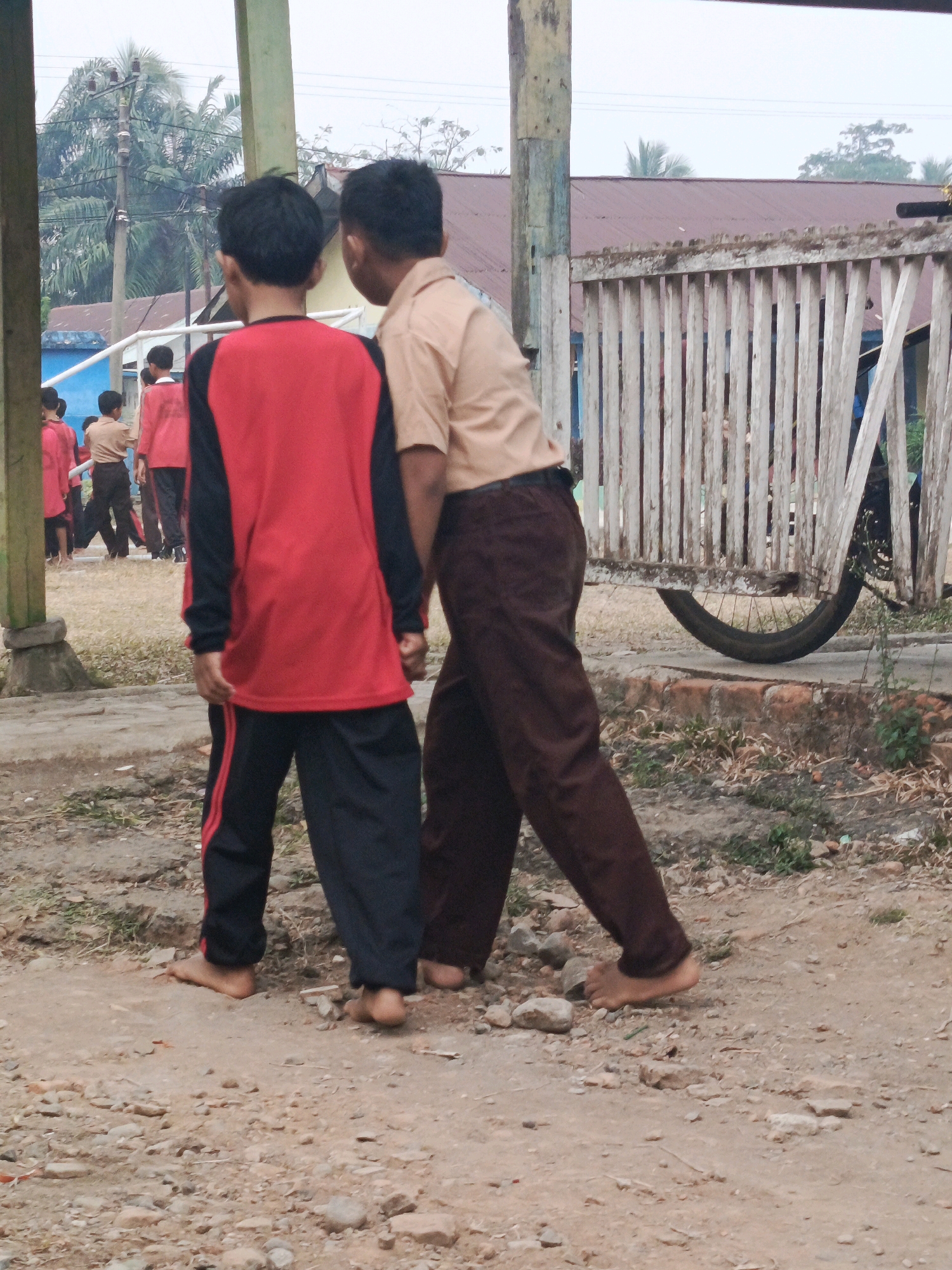 The image depicts an outdoor scene in Megang Sakti II, Indonesia, with two young boys as the central subjects. The boy in the foreground, facing away from the camera, wears a red long-sleeved shirt with black accents and black athletic pants with red stripes. He is barefoot. The second boy, also facing away from the camera but slightly to the right, is wearing a light brown collared shirt and brown pants. He is also barefoot. They appear to be interacting, possibly walking or engaged in a conversation, with the second boy's right arm resting on the first boy's shoulder.
In the background, other children in similar uniforms can be seen walking on a pathway. A rustic wooden fence, weathered and with vertical slats, runs across the middle ground. A bicycle wheel is visible behind the fence, suggesting the presence of a bicycle or cart. Beyond the fence, a building with a reddish-brown roof is partially visible, surrounded by greenery. The ground is a mixture of dirt, gravel, and sparse vegetation. The overall atmosphere appears to be that of a schoolyard or a village area. The sky is overcast.