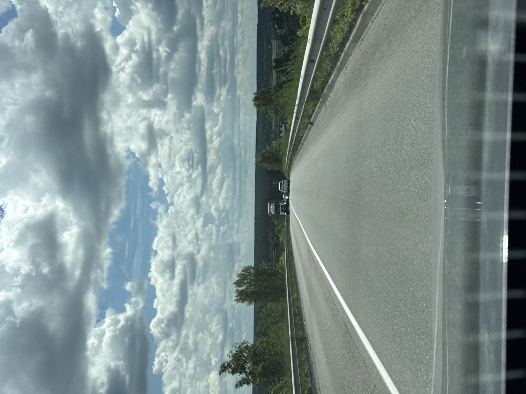 The image shows a view from a car on a paved road with a white line marking the lane. The road curves to the right and slopes slightly uphill. On the left side of the road, there is a metal guardrail. On the right side, there is a grassy embankment with bushes and trees. In the distance, there is a line of trees and a landscape with some buildings. The sky is partly cloudy with a mix of white and grey clouds. Two vehicles are visible on the road ahead: a white car and a white truck. The scene is set in a rural or suburban area, likely in or around Vorzel, Ukraine, given the context provided. The overall impression is a clear, daytime scene on a highway.