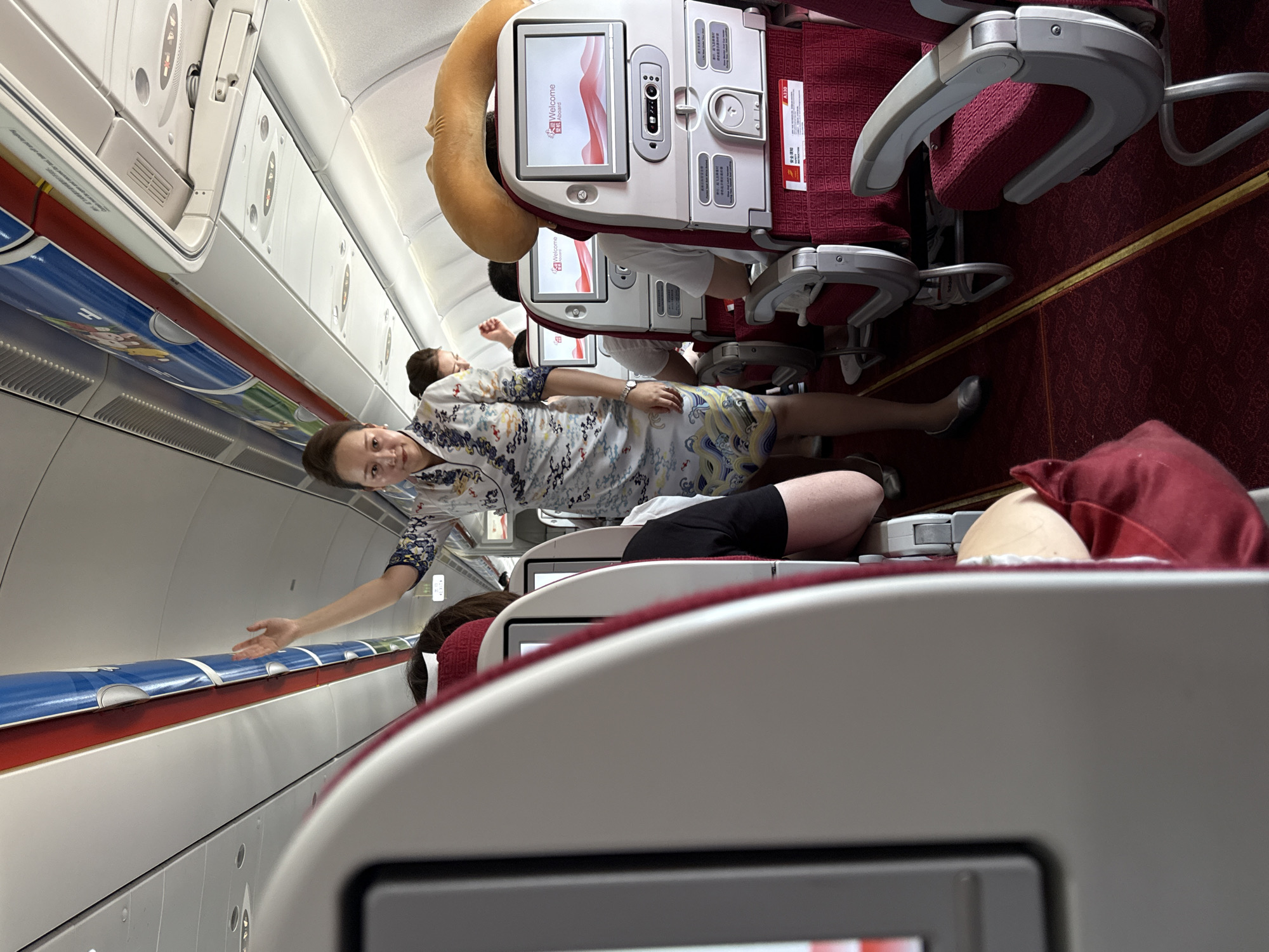 The image depicts the interior of an airplane cabin, likely in the economy or premium economy section. A flight attendant, dressed in a patterned uniform resembling a qipao with blue and white swirling designs and gold accents, stands in the aisle, raising her right hand as if to wave or gesture. She is facing towards the front of the plane and looking slightly to her left. Behind her, other passengers are seated in rows of red upholstered seats with light gray armrests and individual entertainment screens. The aisle is carpeted in red with a yellow stripe. Overhead compartments are visible, one with a blue and white decorative strip featuring cartoon characters. The seats have safety information cards and screens displaying a "Welcome Aboard" message. A U-shaped travel pillow is visible on one of the seats. The lighting suggests daytime. The location is stated as the city of Xiaoshan District, China, implying this is likely a flight departing from or arriving at Xiaoshan International Airport.