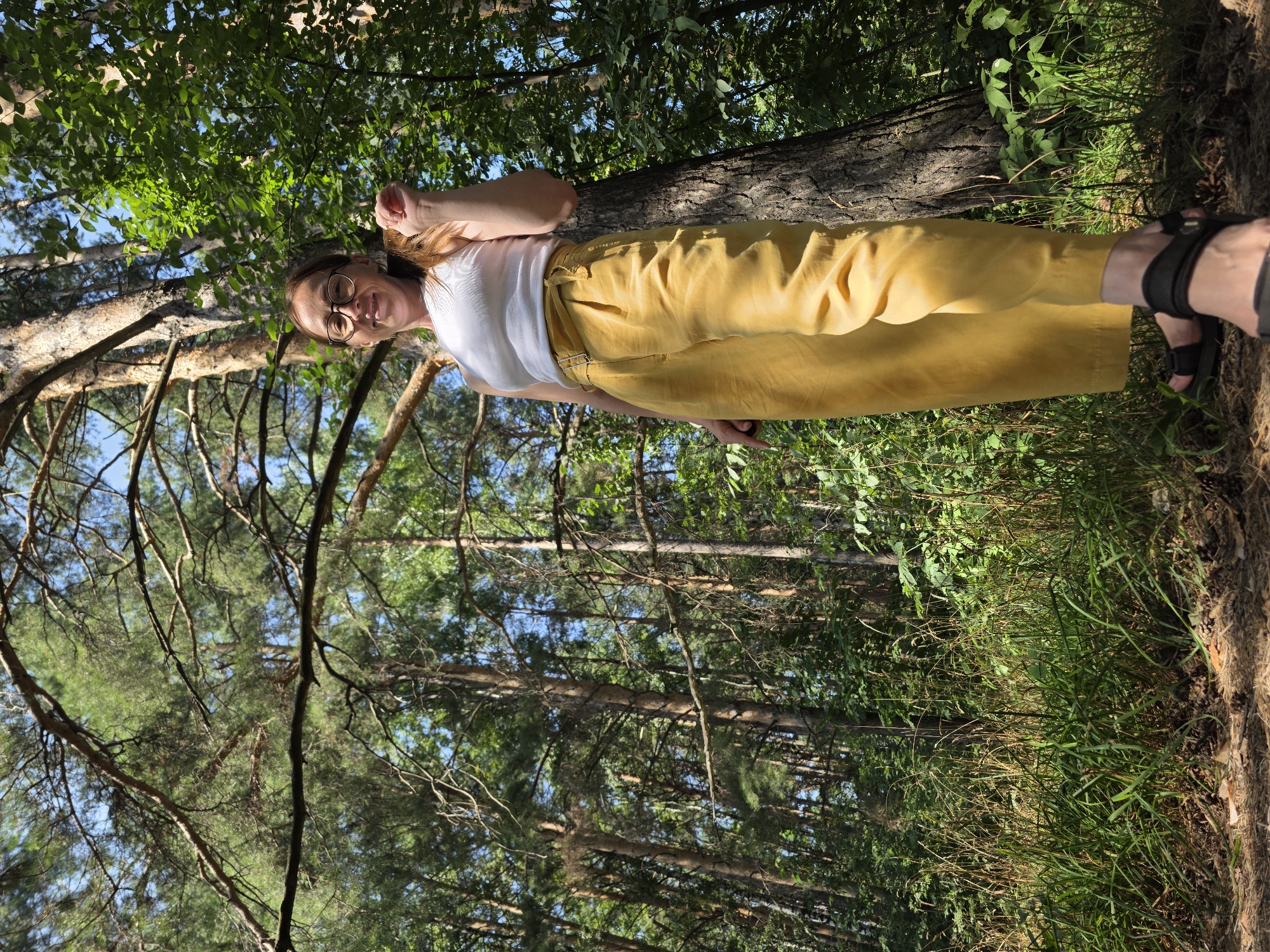 A low-angle shot shows a woman standing in a forest. She is wearing a white tank top and mustard-colored pants. The woman has light brown hair and glasses. She is looking at the camera and smiling. The forest has tall pine trees and green foliage. The ground is covered in pine needles and pine cones. The image appears to have been taken in a natural area, possibly a park or nature reserve. There is no specific indication of the city of Chaikovsky, Russia.
