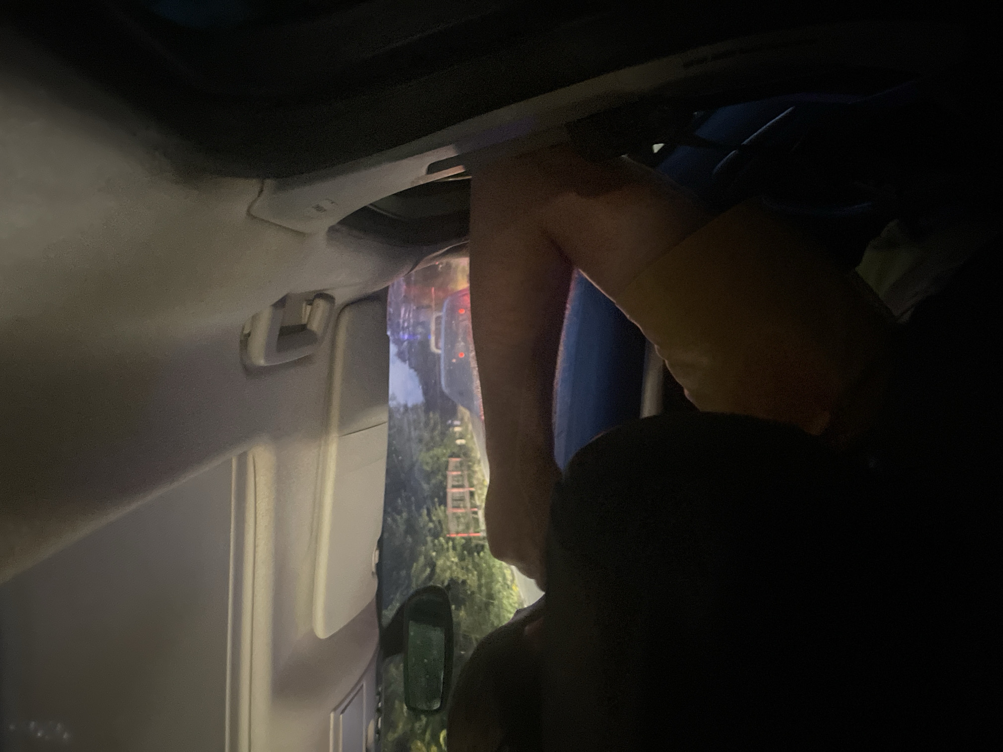 The image shows a person's legs and torso inside a car, with the car's interior visible in the foreground. The person is wearing tan shorts. Outside the car, through the windshield, trees and a red ladder are visible. The lighting suggests it is dusk or nighttime, with artificial lights from what appears to be a street or another vehicle reflecting on the car's interior and windshield. The scene appears to be in a somewhat rural or suburban setting, given the trees and ladder. The angle of the photo is unusual, pointing upwards and outwards from inside the car, making it difficult to discern the exact activity taking place. No text is visible in the image. The location context provided is Beloci, Moldova.