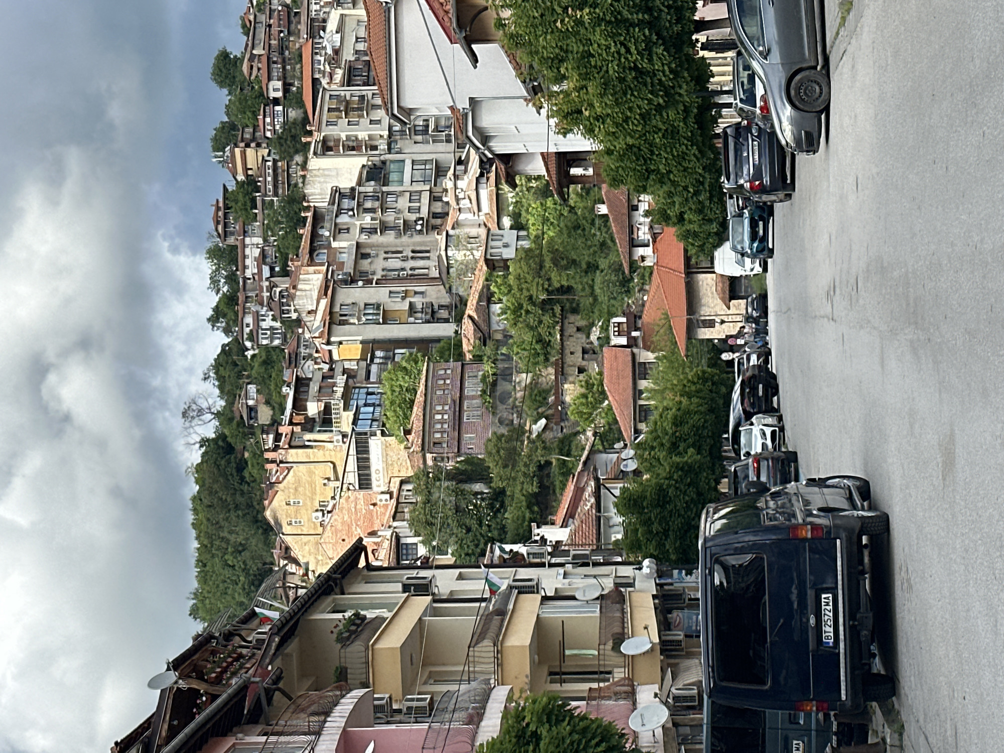 The image depicts a street scene in Veliko Tarnovo, Bulgaria. The street slopes downwards, with cars parked along the right side. A dark blue van is prominent in the foreground, displaying a license plate with "BT 2572 MA." A row of other parked cars extends into the distance. The left side of the frame showcases a dense urban landscape, with multi-story buildings cascading down a hillside. These buildings vary in color and architectural style. Satellite dishes are visible on some of the buildings. There is greenery interspersed among the buildings. The sky is overcast with dark clouds. The Bulgarian flag can be seen near the bottom left.