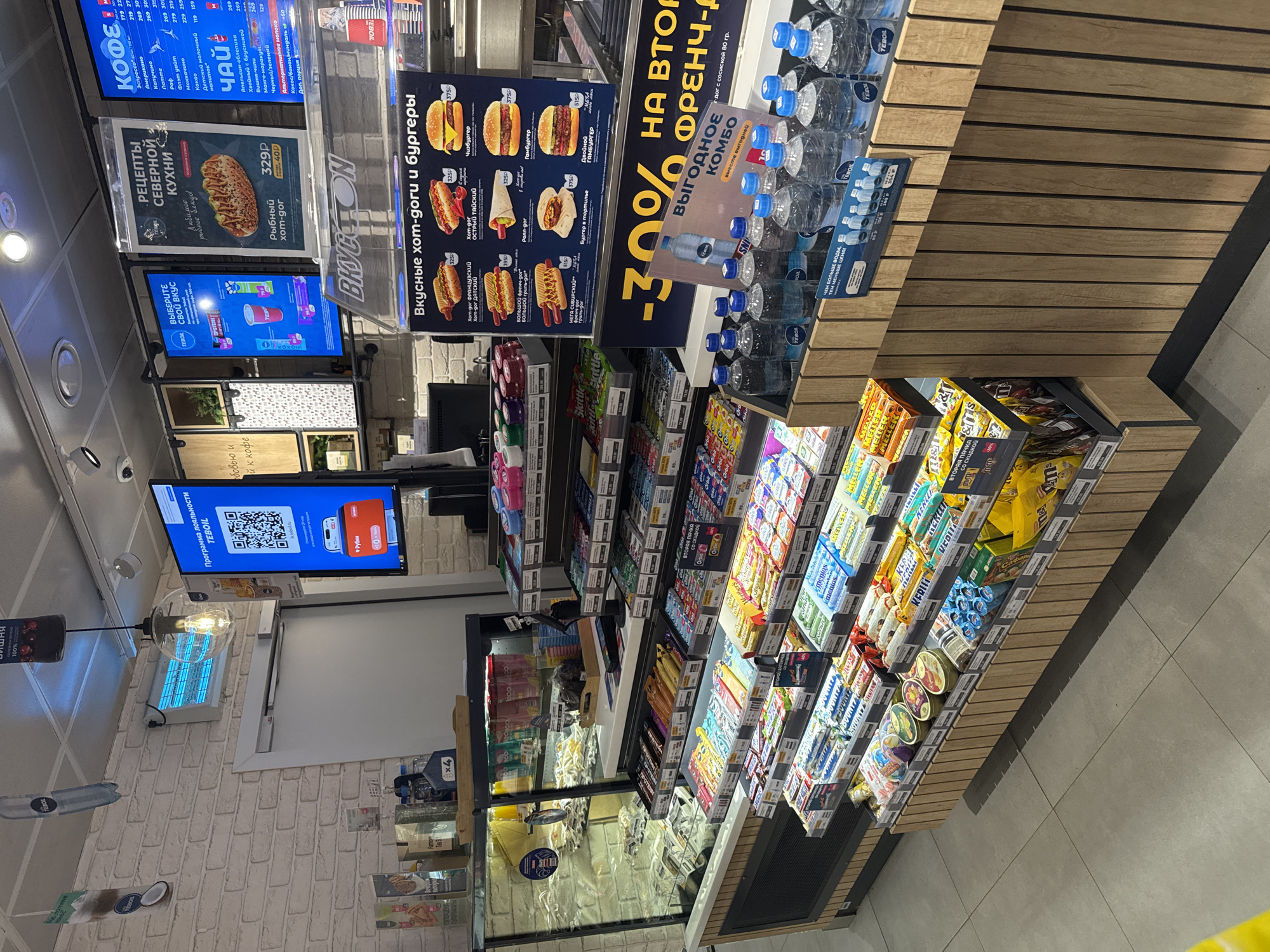 The image depicts an interior view of a convenience store in Saint Petersburg, Russia. Signage in Russian advertises a variety of food and drink options including coffee, tea, hot dogs, and burgers. A promotional sign highlights a 30% discount on a second french hot dog when purchased as a combo. Shelves are stocked with bottled water, candy bars, and other snacks. A digital display shows a QR code for a loyalty program. The environment is well-lit and appears clean.