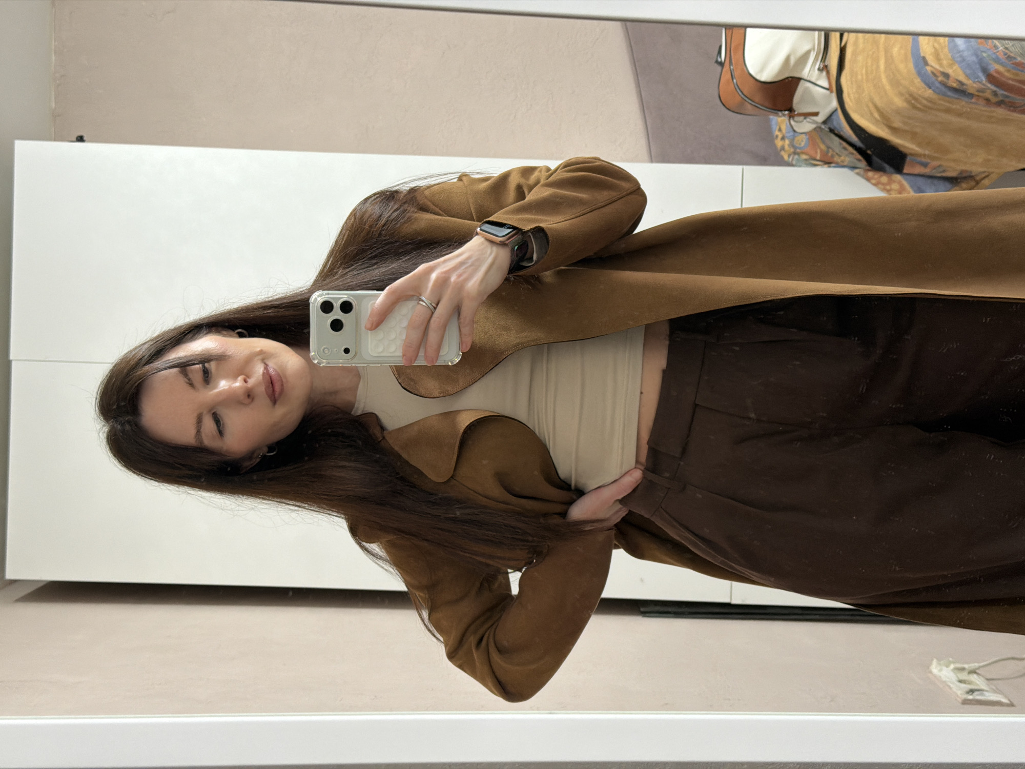 A woman stands in front of a mirror, holding a smartphone to take a selfie. She is wearing a brown coat, beige top, and brown trousers. The background features a white wall with what looks like a door or a closet. The location appears to be indoors. The presence of a mirror and personal clothing suggests it might be a private residence. There is no indication in the image that it is located in Perm, Russia.