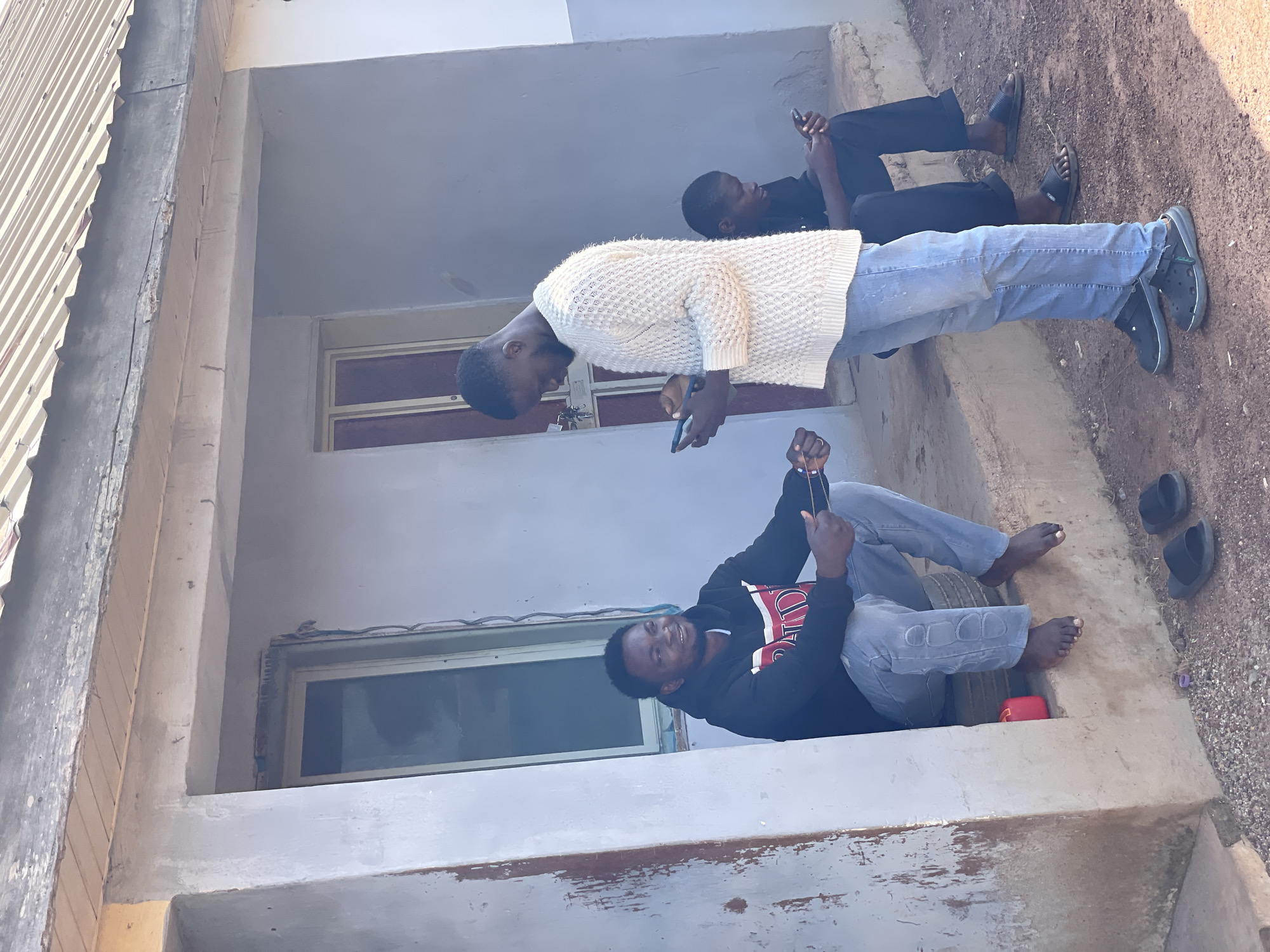 The image depicts three young men outside a building in what appears to be a residential area in Durumi, Nigeria. The building has a light gray exterior and a corrugated metal roof. Two men are seated on the concrete steps leading to the entrance, while one man stands.
The man on the far left, seated on the steps, is wearing a black hooded sweatshirt with red and white stripes on the chest and light blue jeans. He is looking to his right and smiling, holding a string or wire in his hands. He is barefoot.
The standing man in the center is wearing a textured white long-sleeved sweater and light blue jeans. He is holding a smartphone in his left hand and looking down at it. He is wearing dark-colored clogs on his feet.
The third man, on the far right, is seated on the steps behind the standing man. He is wearing a black t-shirt and black pants, and is looking down at a smartphone in his hands. He is wearing black sandals.
The ground in front of the steps is covered with dirt and small stones. Two black flip-flops are visible on the ground near the standing man's feet. A red cylindrical object is partially visible on the steps near the seated man on the left. The overall scene suggests a casual, outdoor gathering.