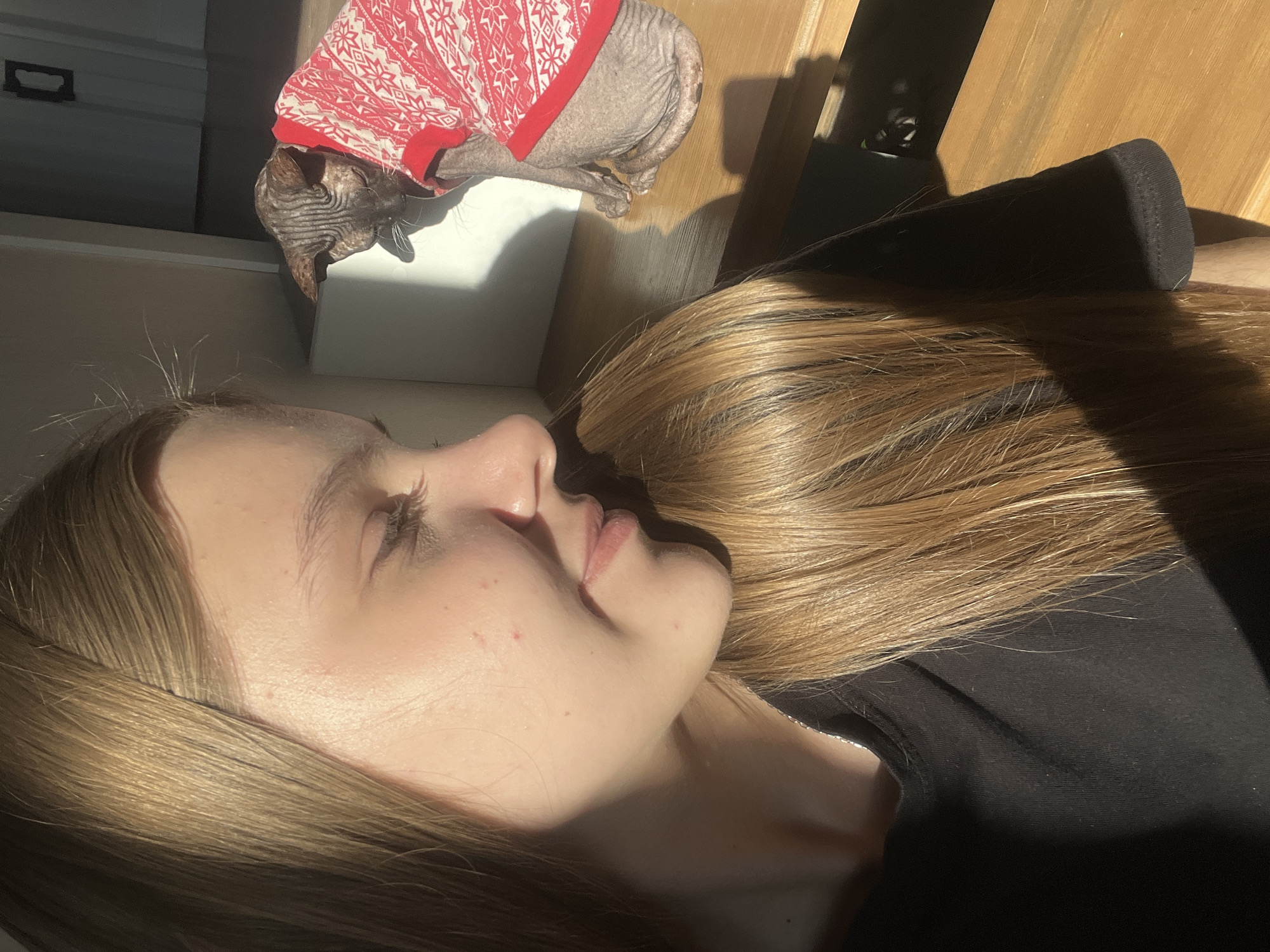 A close-up photograph depicts a light-skinned person with blonde hair, lying down, likely indoors, as suggested by the controlled lighting and presence of furniture. The person is wearing a black top. Beside the person is a hairless cat wearing a red and white sweater. The cat is lying on top of a white object. The setting seems to be a domestic environment. There is no information to suggest that this image was taken in Votkinsk, Russia.
