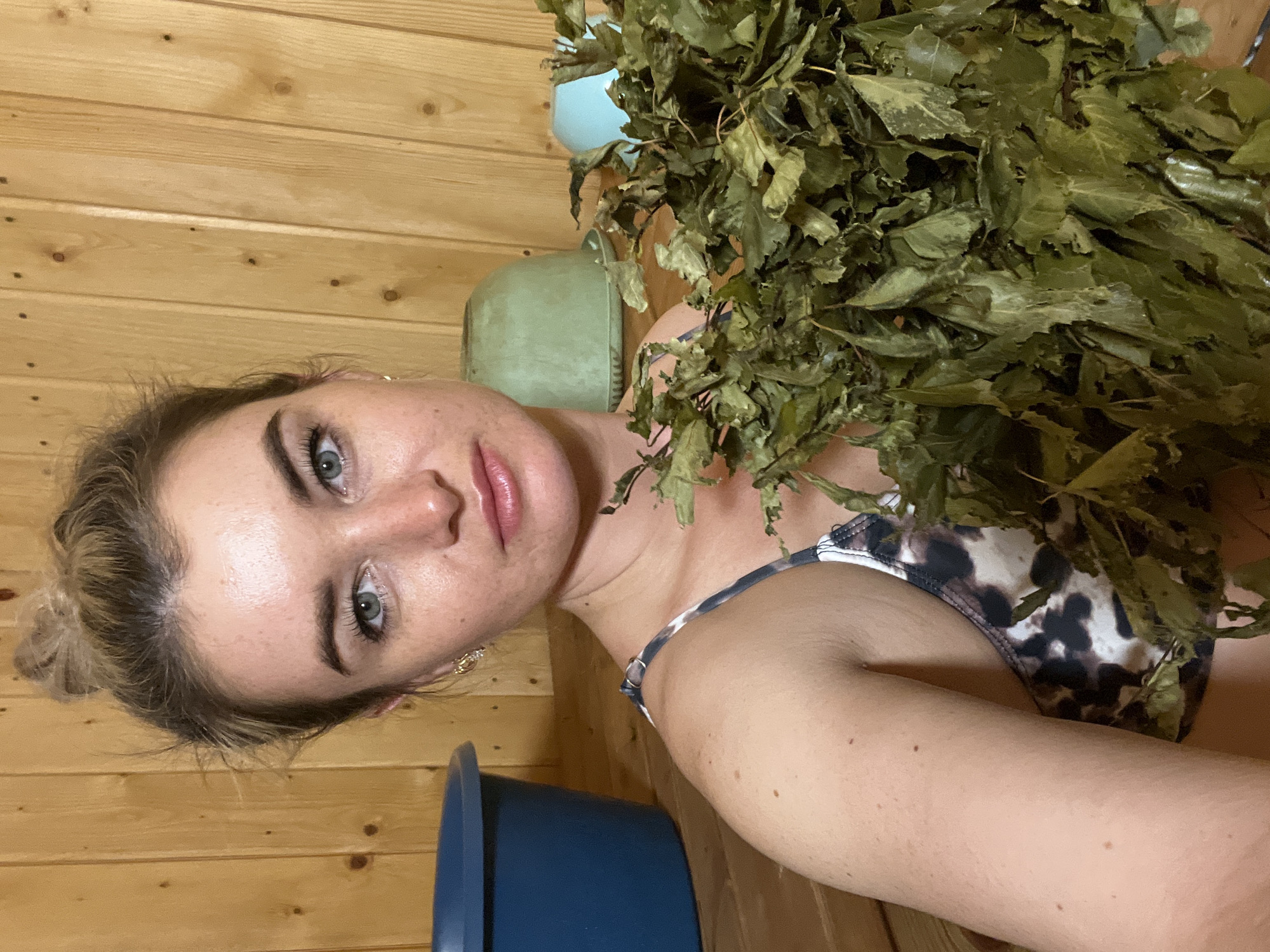 A woman with fair skin and blonde hair pulled back in a bun is holding a bundle of dry green leaves while standing in a sauna made of light-colored wood. She wears a spotted swimsuit. A blue bucket is visible in the lower left corner, and green buckets are visible in the background. The image is taken at eye level and appears to be a selfie. The location is Гаревая, Russia.