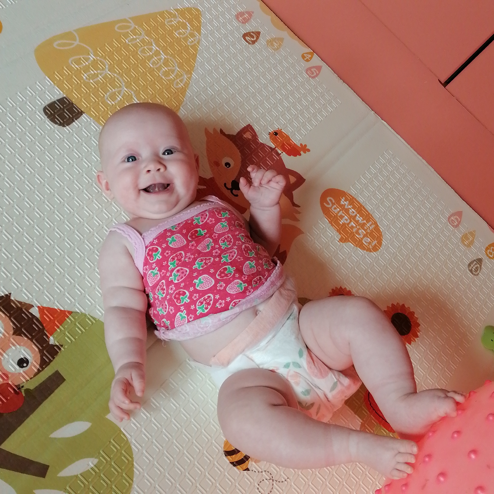 The image shows a baby lying on its back on a play mat. The baby is wearing a pink shirt with a strawberry pattern and a diaper. The baby is smiling and looking at the camera. The play mat has various animal and nature-themed designs, including an owl, a fox, bees, and sunflowers. The background appears to be a room with pink furniture. The image does not contain any information that would allow for its location to be specified within the city of Trubacheyevka, Russia.