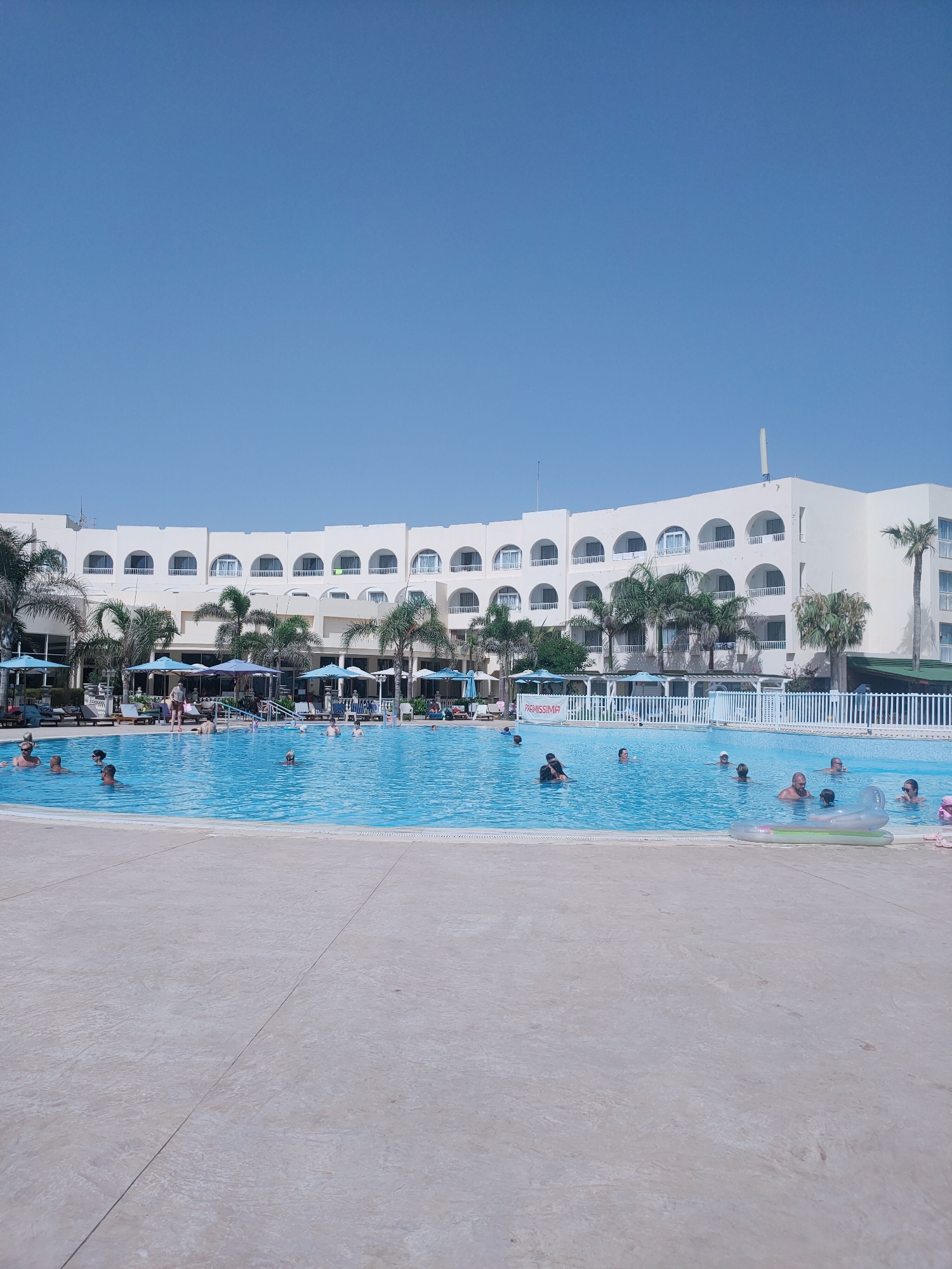 A wide-angle shot captures a bright, sunny day at a resort pool in Nabeul, Tunisia. The foreground is dominated by a large, pale stone patio, leading to a vibrant blue swimming pool filled with people. The pool is oval-shaped, with a white border around its edge. Several individuals are swimming, some with their heads above water, while others are partially submerged. One person is floating on an inflatable mattress, and another is visible on a pink inflatable ring.
Beyond the pool, a row of palm trees and lush green foliage line the edge, interspersed with blue umbrellas providing shade over empty lounge chairs. In the background, a long, multi-story, white hotel building with arched windows and balconies curves across the scene. A few more palm trees are scattered around the hotel. The sky is a clear, cloudless blue. The overall impression is one of leisure and vacationing in a warm climate.