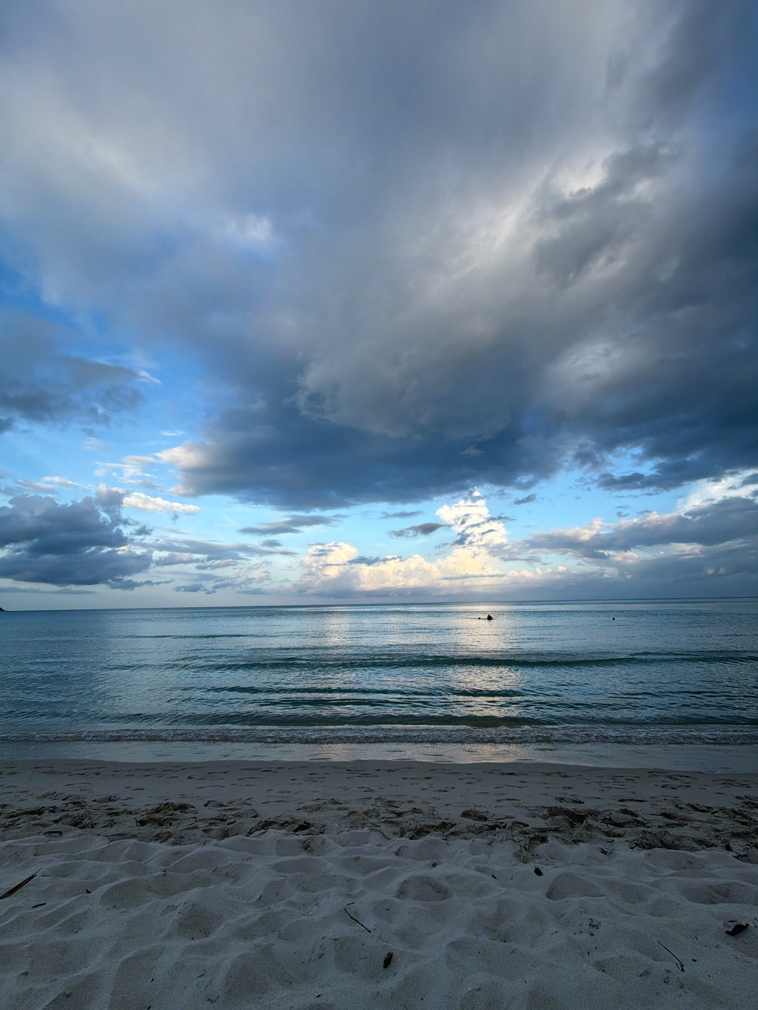 A wide-angle, low-angle shot captures a beach scene in Ko Samui District, Thailand. The foreground is dominated by white sand with footprints and some scattered debris. Gentle waves lap at the shore, creating horizontal lines on the turquoise water. In the distance, a person is visible in the water, appearing to be swimming or surfing. The horizon is marked by the ocean meeting a dramatic sky filled with dark, heavy clouds interspersed with patches of blue. The overall atmosphere suggests an overcast day, possibly transitioning to or from a storm.