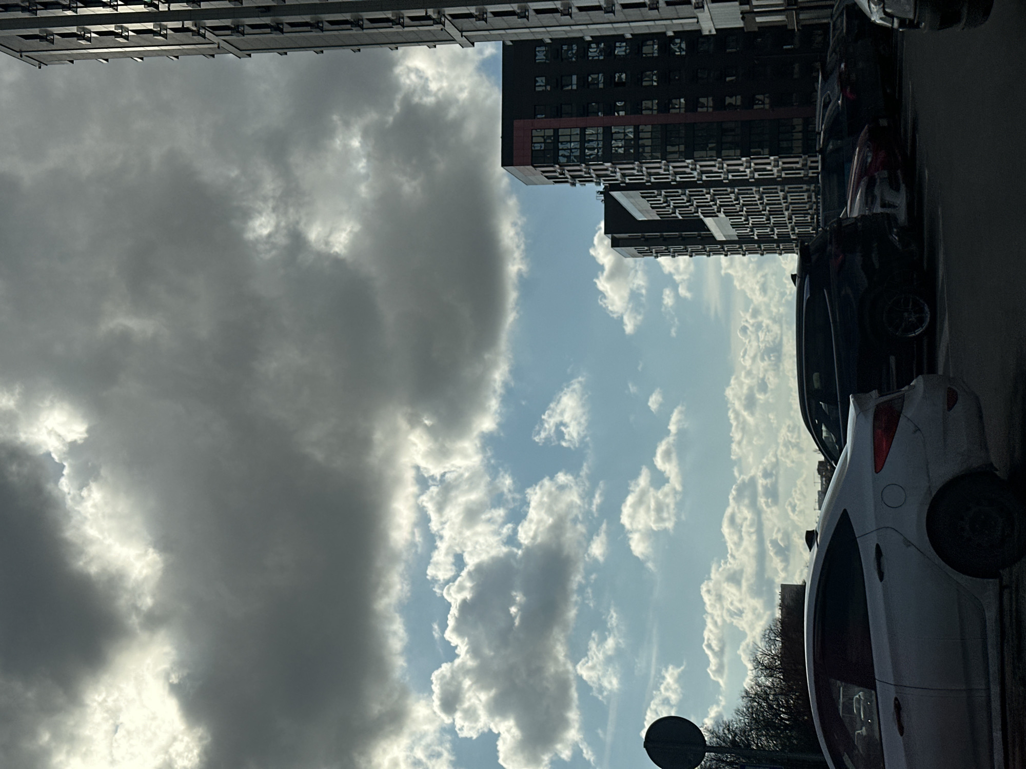 The image depicts a street scene in Rostov-on-Don, Russia, under a partly cloudy sky. High-rise residential buildings flank the sky, with one building showing a facade pattern of dark windows and light balconies. Several parked cars line the right side of the frame. A white car is closest to the viewer. A traffic sign is visible in the lower portion of the scene, near bare trees. The sky features a mix of dark, overcast clouds and patches of blue with white, fluffy clouds.