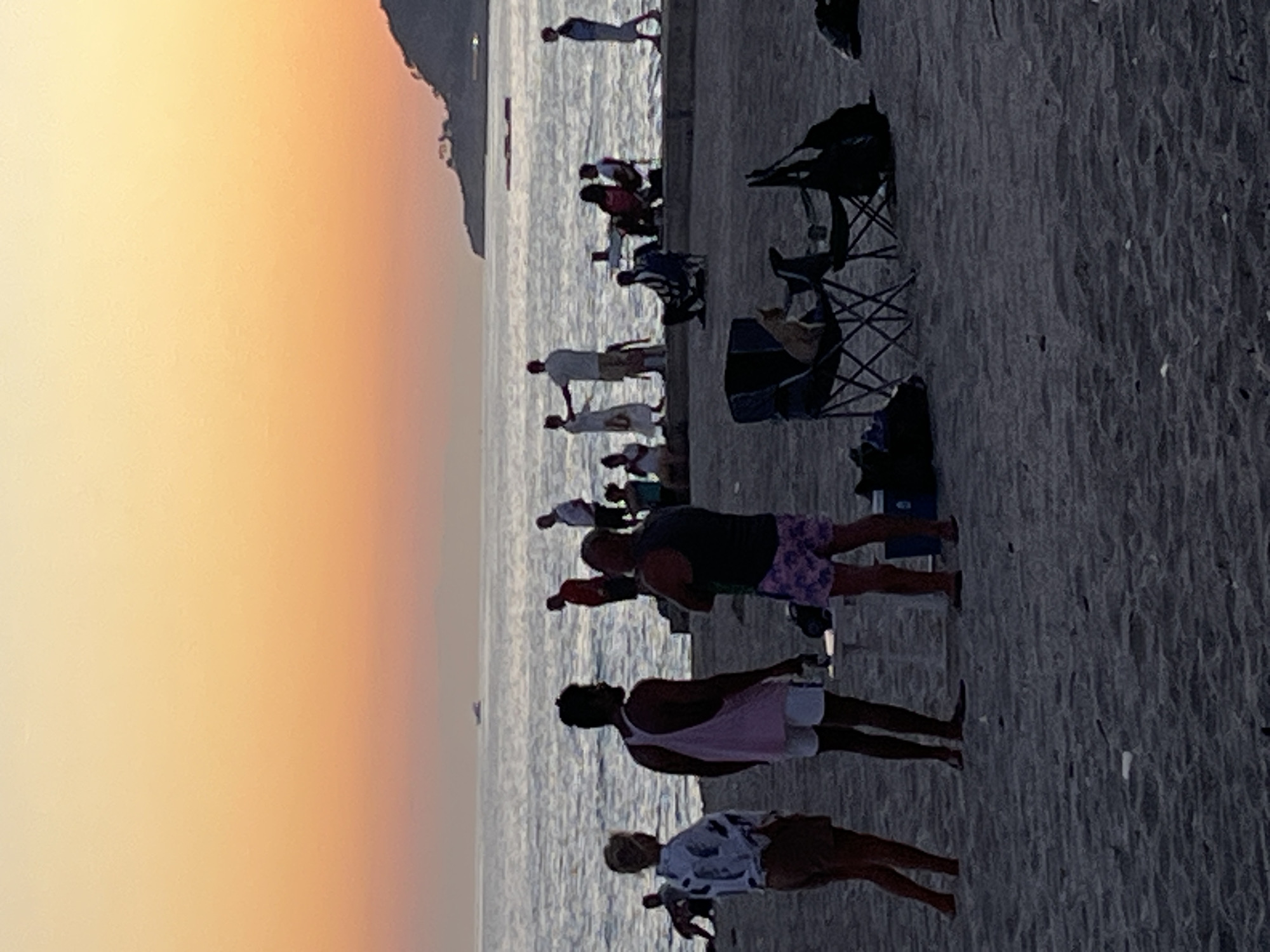 The image depicts a beach scene at sunset in Alanya, Turkey. Several people are visible along the shoreline. Some are standing, while others are seated in chairs near the water's edge. The sky transitions from a bright yellow to orange as the sun sets over the sea. A land mass is faintly visible in the distance.