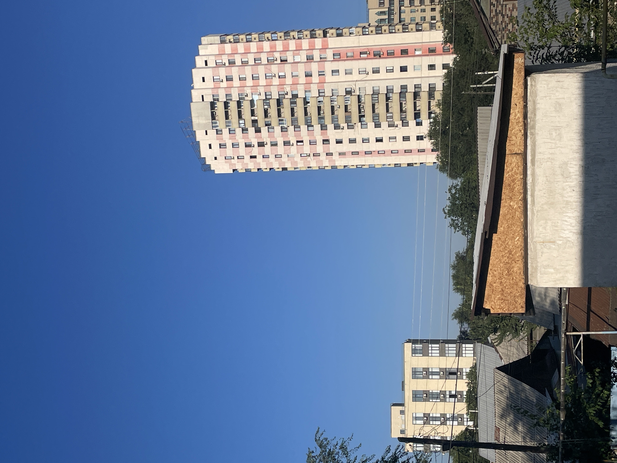 The image depicts an urban scene under a clear blue sky. In the foreground, there are several low-lying structures, some with corrugated metal roofs, and a building under construction with a plywood-covered upper section. In the mid-ground, behind these structures and a line of trees, stands a light-yellow rectangular building with multiple windows. Further in the background, a tall, multi-story apartment building with a cream and pink facade dominates the right side of the frame. The apartment building features numerous balconies and windows, and a section of what appears to be scaffolding or construction equipment is visible on its upper edge. The scene is characteristic of residential areas in Almaty, Kazakhstan, with a mix of older and newer construction. No people are visible in the image. The environment appears to be a sunny day.