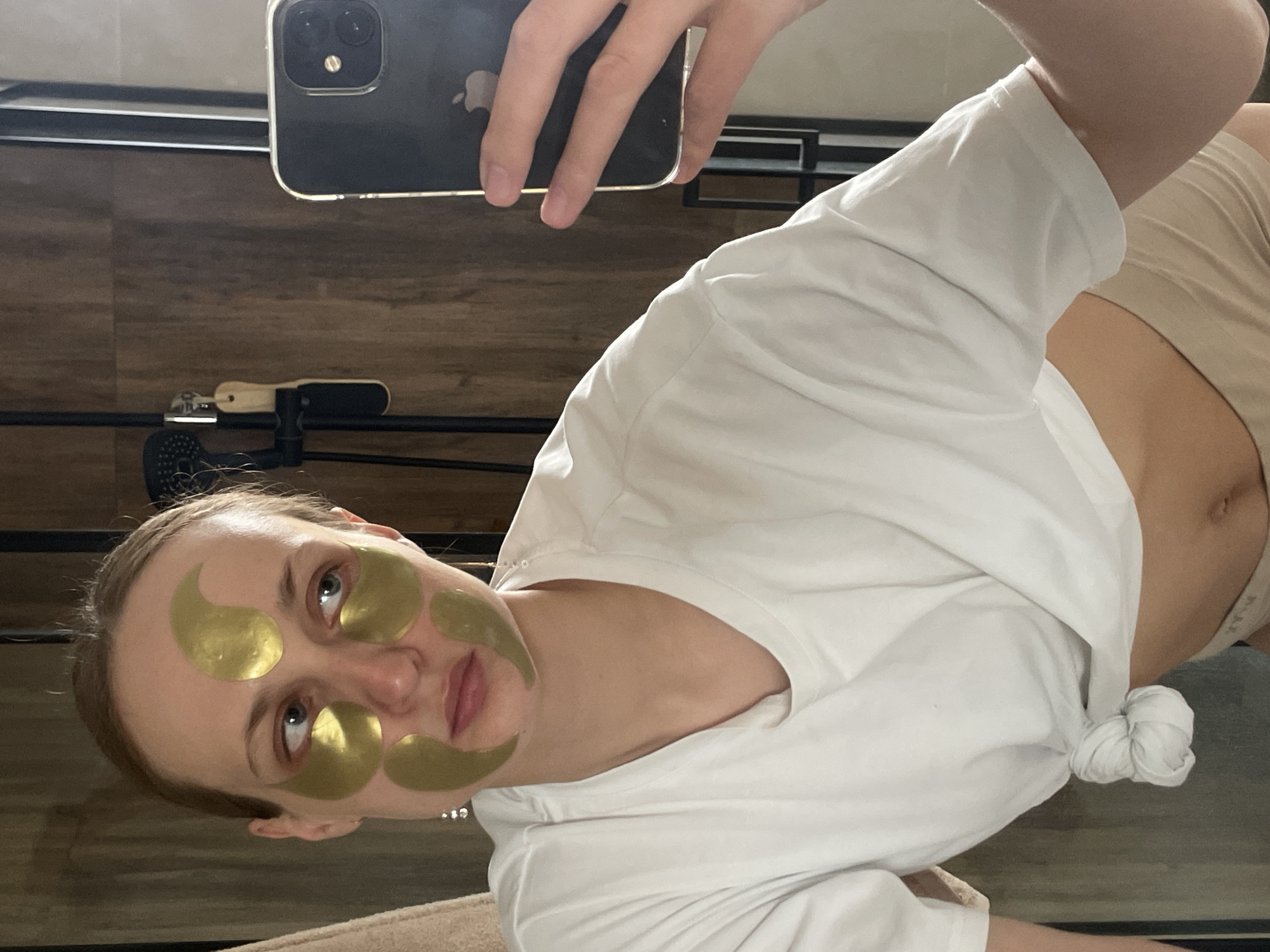 A woman is taking a selfie in what appears to be an indoor setting. She has gold eye and facial patches applied. She is wearing a white t-shirt tied in a knot. Her face is angled towards the camera, with her left eye more visible. She is holding a black smartphone, likely to take the photo. The background includes a wooden surface, and a black metal structure with a shower head. Location data suggests this is in Подшивалово, Russia.