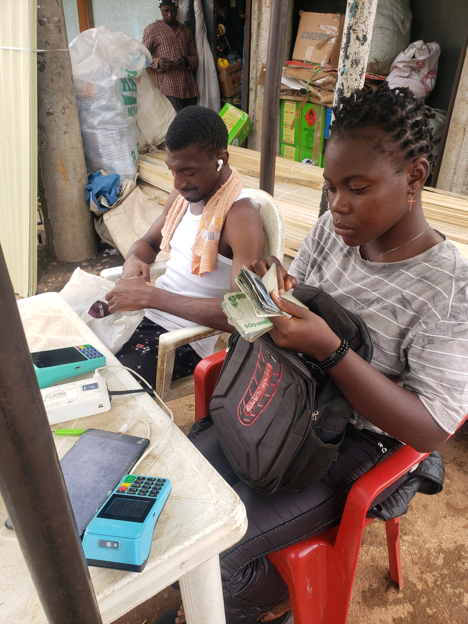 In Sabon Tasha, Nigeria, two individuals are engaged in a transaction at a table. A man, wearing a white tank top and an orange towel draped over his shoulder, is seated, looking down and holding a white plastic bag. Next to him, a woman is seated, wearing a grey and white striped t-shirt and black pants. She is counting a stack of banknotes, with a black backpack resting on her lap. On the table in front of them are various electronic devices, including a blue point-of-sale terminal and a white power bank. In the background, stacks of lumber and bags are visible, suggesting a storage or marketplace environment. A man in a plaid shirt is standing in the background, observing.