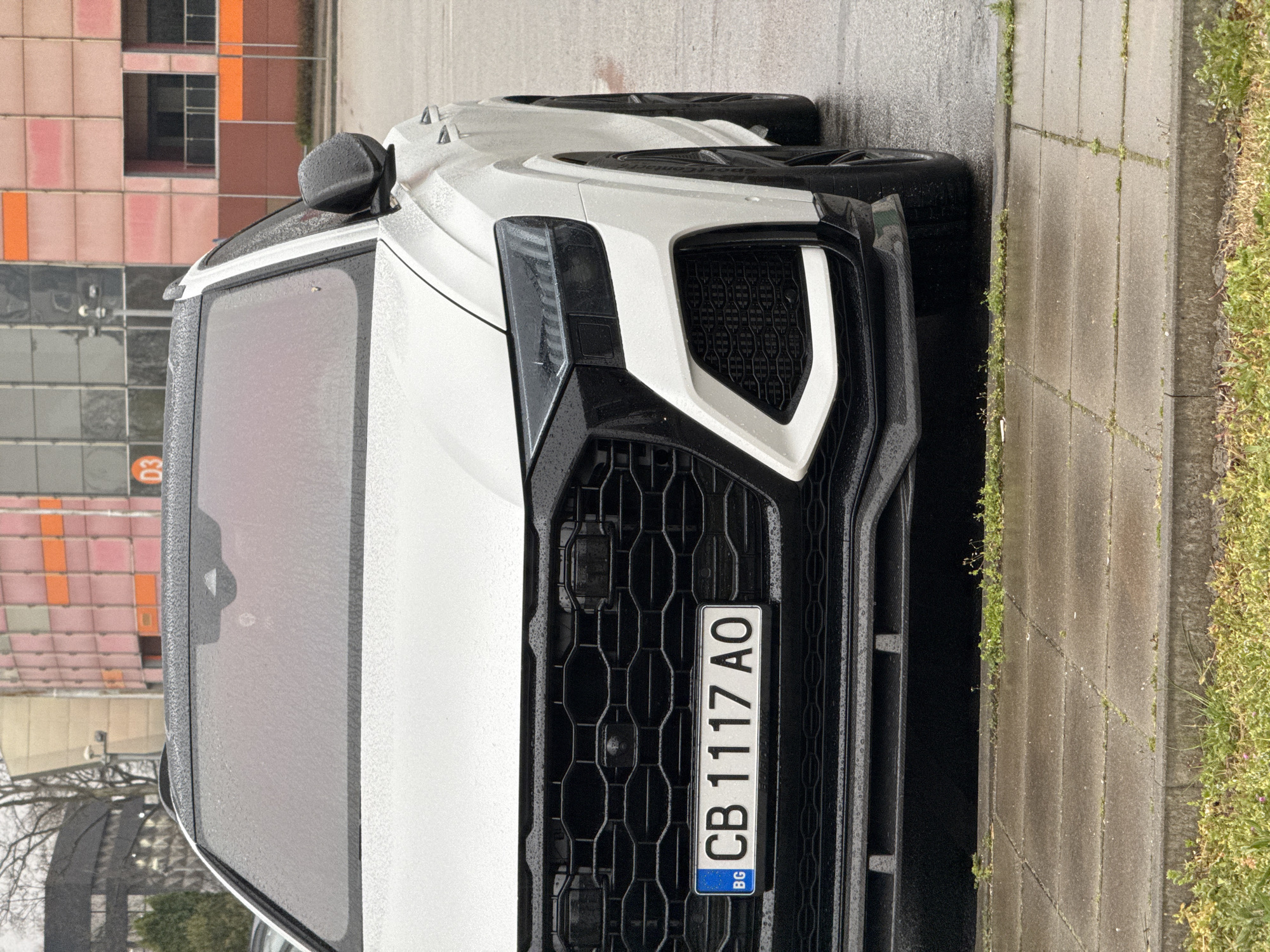 A medium shot depicts the front of a white car parked on a wet street, likely in Plovdiv, Bulgaria. The car's grill is prominently featured, displaying a Bulgarian license plate reading "CB 1117 AO." Water droplets cover the car's surface, suggesting recent rain. The background shows a multi-story building with a mix of peach and orange colored sections. A narrow sidewalk is visible beside the car, with grass growing along the edge. The overall scene conveys a rainy day in an urban environment.