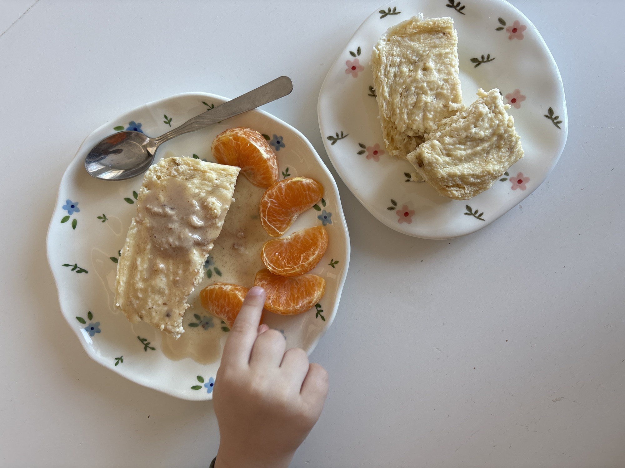 The image shows two floral-patterned plates on a white surface. The plate on the left contains a rectangular piece of food covered in a creamy sauce, mandarin orange slices, and a spoon. A child's hand reaches towards the orange slices. The plate on the right holds two pieces of similar food without sauce.
There is no evidence of the location in the city of Ulyanovsk, Russia.