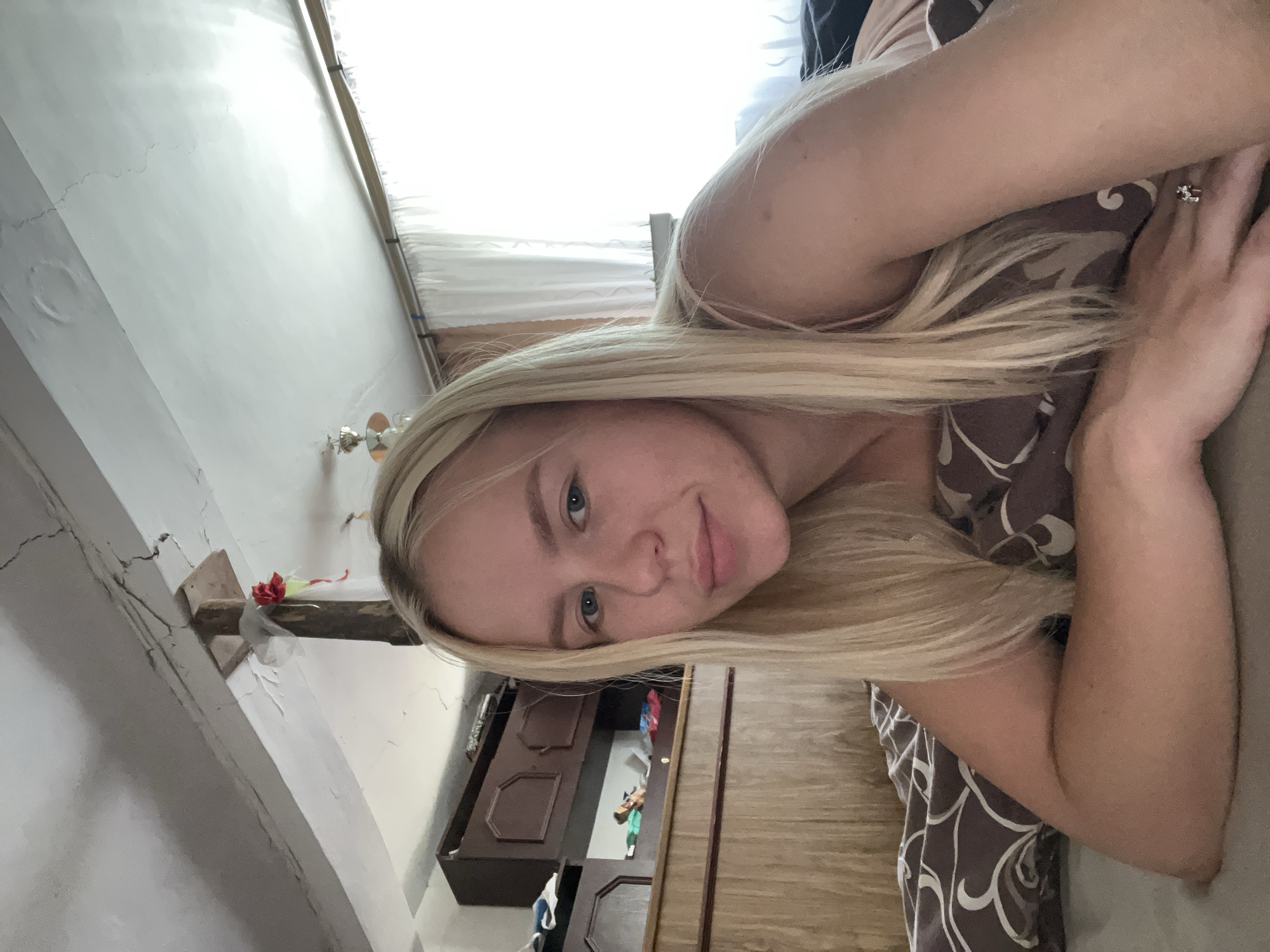 The media file is a photograph of a young woman with blonde hair, lying on her side and looking towards the camera. She has blue eyes and is wearing a patterned top. Her left arm is resting on her chest, and she is wearing a ring on her left hand. The background appears to be a room with white walls, a wooden cabinet, and a window with white curtains. There is a small red decoration with white netting on a wooden beam in the background. The lighting suggests it might be daytime, possibly indoors with natural light coming from the window. The woman's expression is neutral. The photograph is taken from a slightly elevated angle. The image is oriented sideways.