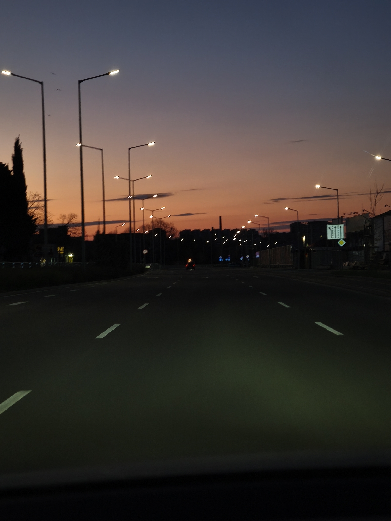 The image shows a road at night in Burgas, Bulgaria, illuminated by streetlights. The sky displays a gradient of colors from orange at the horizon to dark blue above. Streetlights line both sides of the road, extending into the distance. Road markings are visible on the asphalt. On the right side of the road, a road sign is visible. A single car headlight appears in the distance. The photo is taken from within a vehicle.