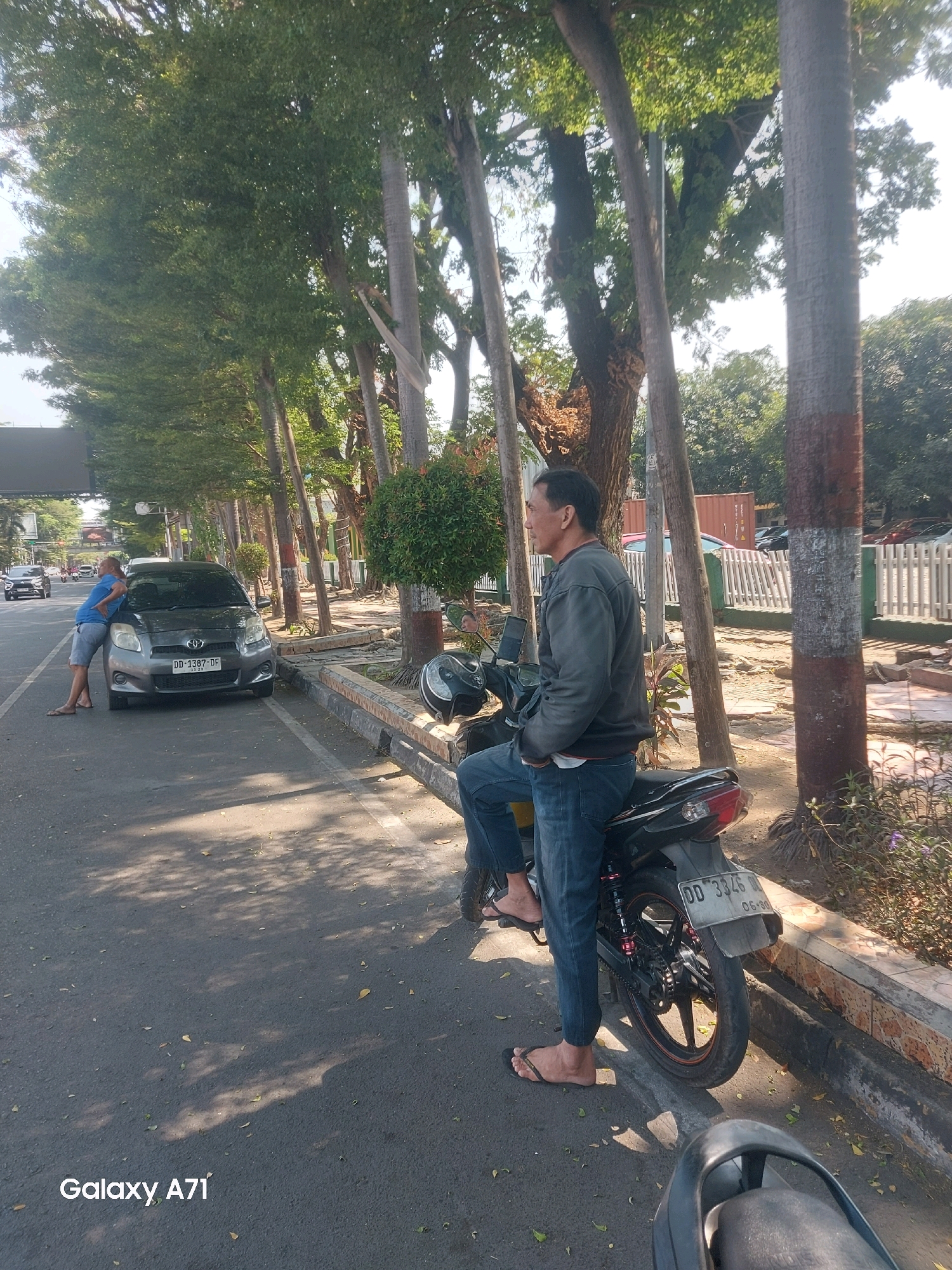 In Makassar, Indonesia, a man in a grey long-sleeved shirt and blue jeans is seated on a black motorcycle parked on the side of a street. He is looking to his left. Another man, wearing a blue t-shirt and grey shorts, is leaning on the hood of a dark grey car parked in front of him. The car has a license plate that reads "DD 1387 DF." Trees line the street, casting shadows on the asphalt. A white picket fence is visible in the background, along with some buildings and a shipping container. The overall scene suggests a typical urban environment on a sunny day.